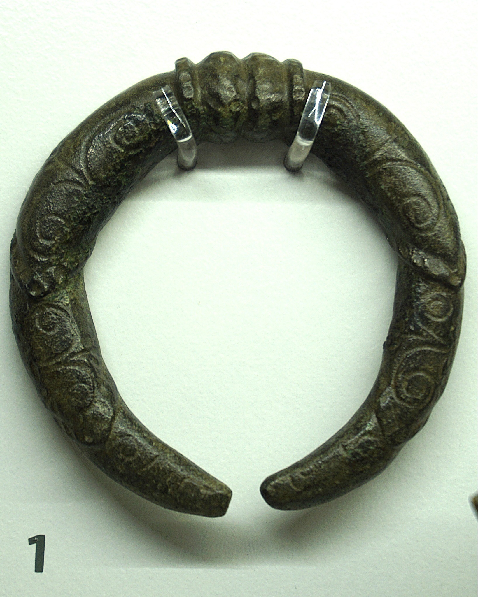 Dieser Alternativtext wurde KI-generiert: Ein dunkler, gebogener Gegenstand mit aufwendigen Verzierungen ist zu sehen. Das Objekt befindet sich vor einem hellen Hintergrund und wird von zwei Metallstreifen gehalten.