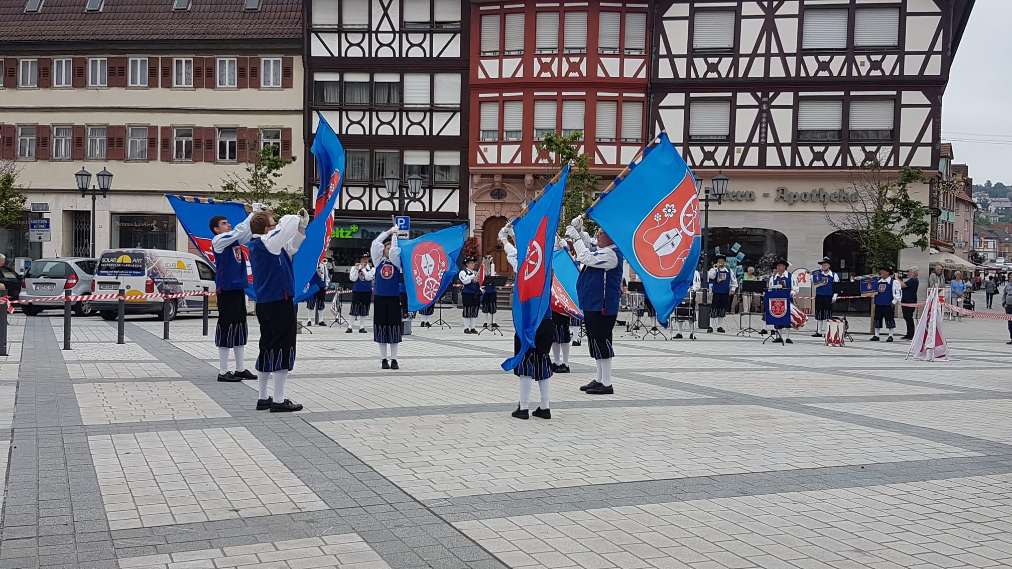 Dieser Alternativtext wurde KI-generiert: Eine Gruppe von Personen in Uniformen hält Fahnen in einer städtischen Umgebung. Im Hintergrund befindet sich ein Fachwerkgebäude und ein Fahrzeug.