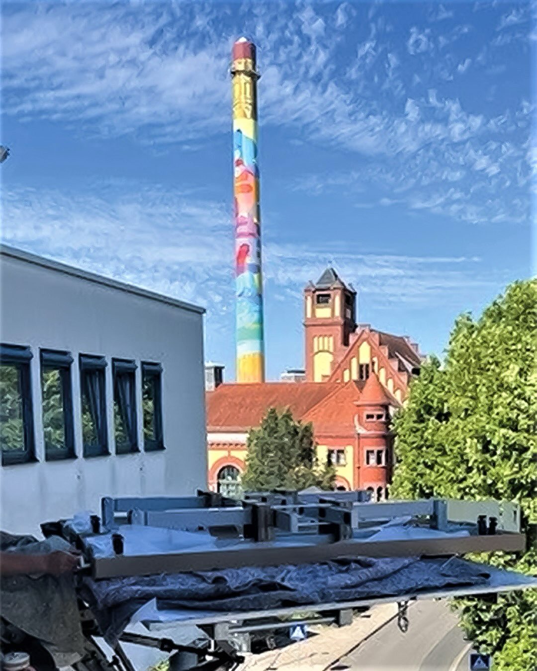Dieser Alternativtext wurde KI-generiert: Ein hoher Kamin mit farbenfroher Bemalung erhebt sich über Gebäude mit roten Ziegeln und Bäumen. Der Blickwinkel ist von einem Balkon oder Dach aus aufgenommen.