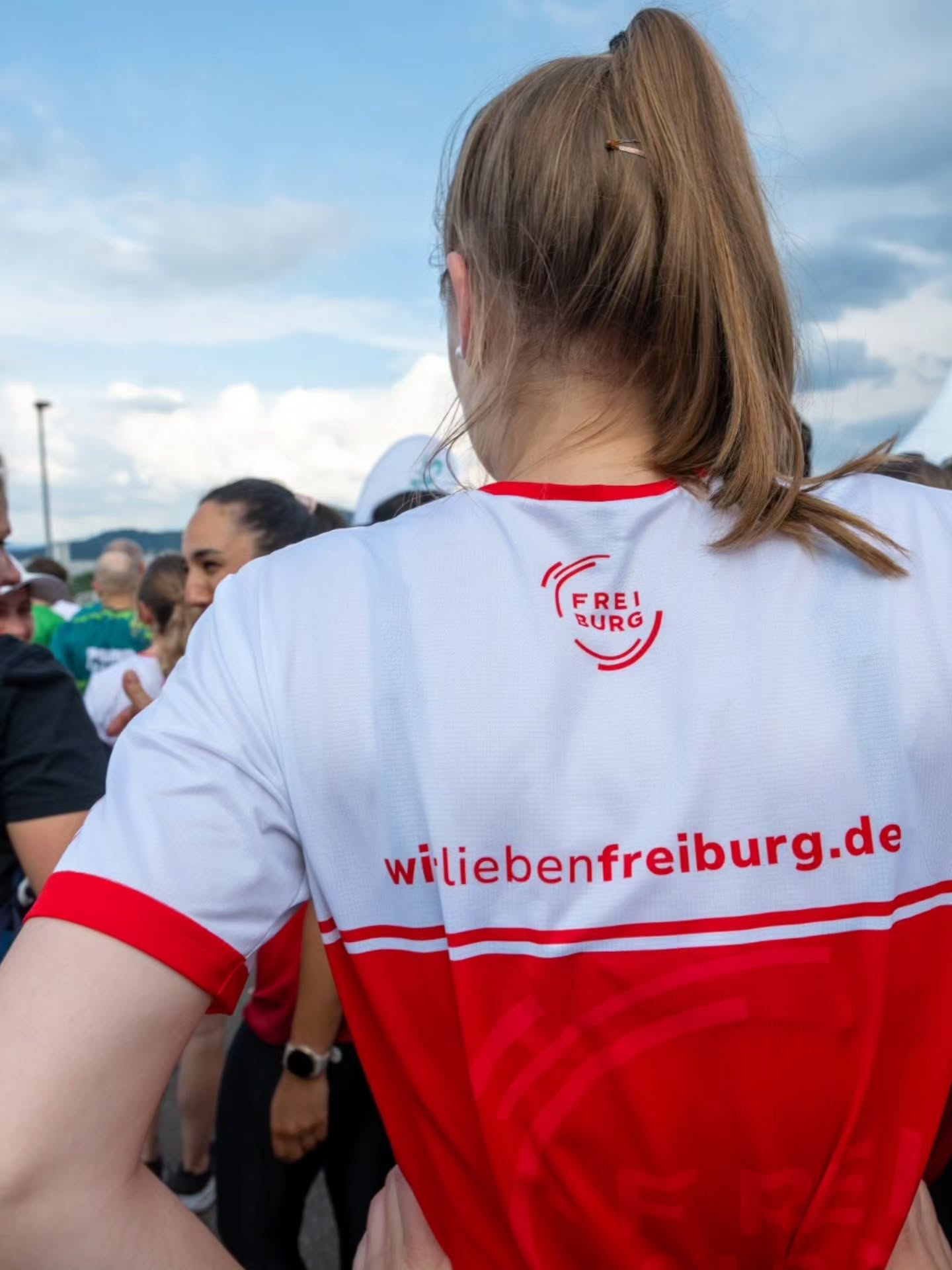 Eine Person mit langem, zu einem Pferdeschwanz gebundenem Haar steht mit dem Rücken zur Kamera und trägt ein weißes T-Shirt mit rotem Saum und rotem unteren Bereich. Auf dem Rücken des Shirts ist ein rotes Logo mit der Aufschrift "FREIBURG" sowie die Webadresse "wiliebenfreiburg.de" zu sehen. Im unscharfen Hintergrund befinden sich weitere Menschen unter einem blauen Himmel mit Wolken. (KI-Alternativtext)