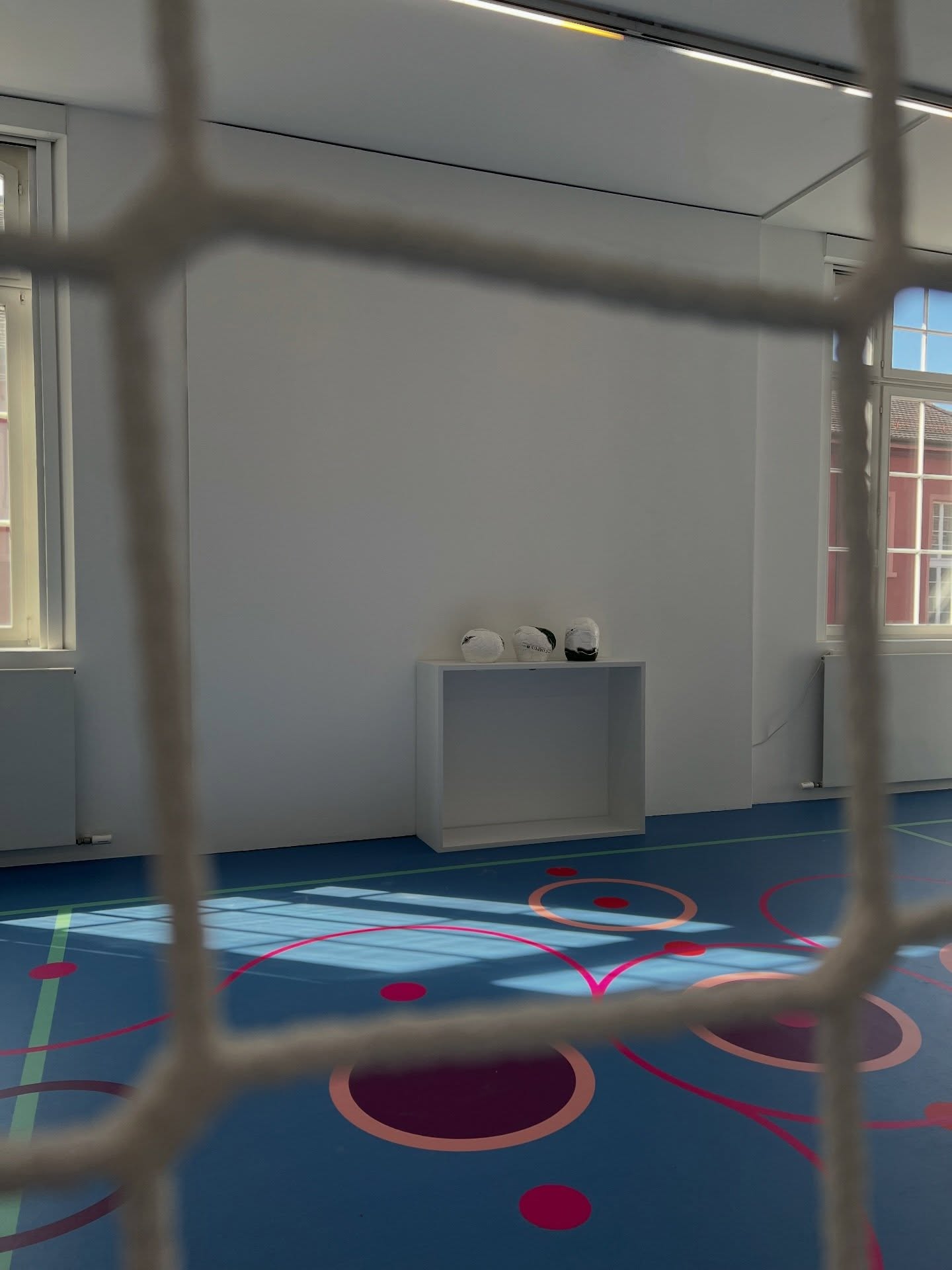 Dieser Alternativtext wurde KI-generiert: Ein Innenraum mit hellen Wänden und einem bunten Boden. Auf einer niedrigen Plattform liegen zwei dunkle Objekte.