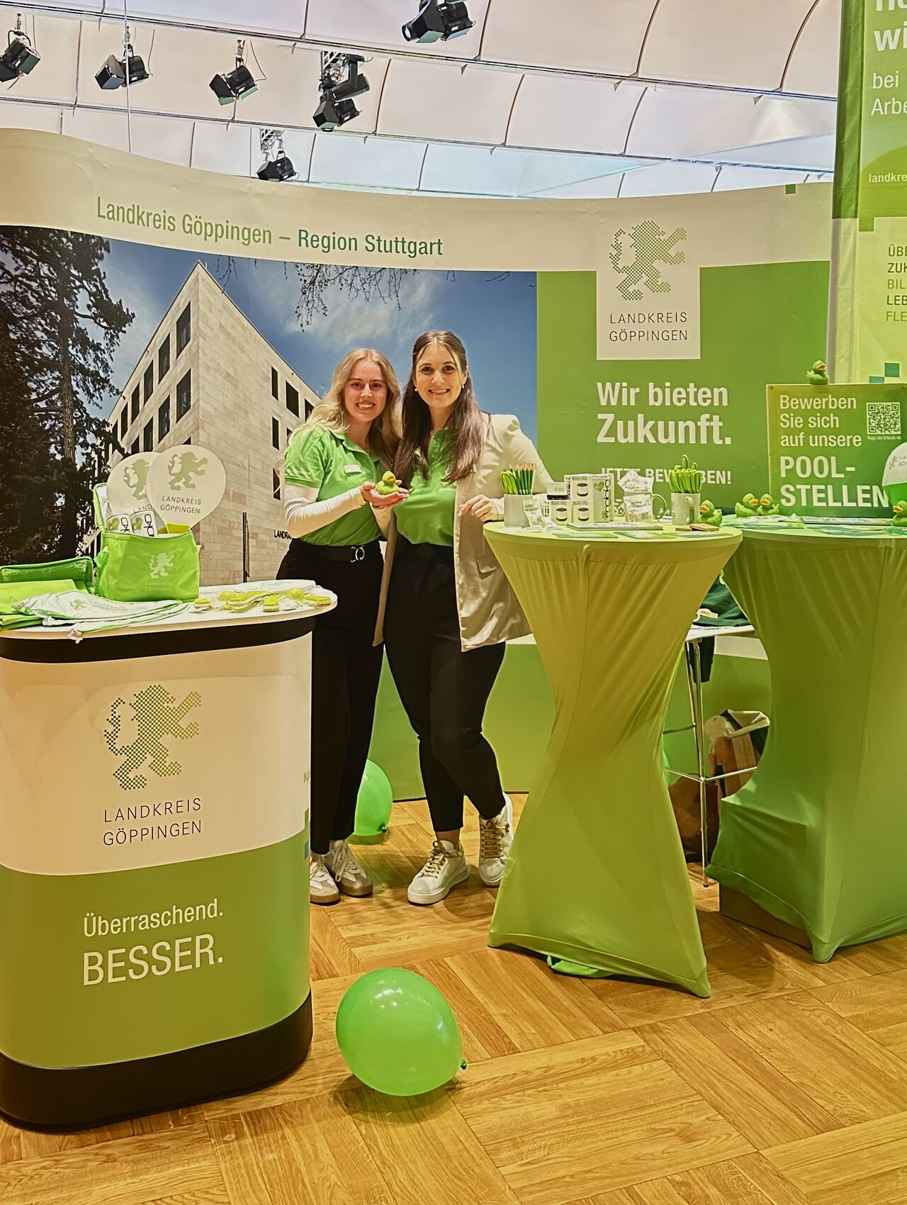 Dieser Alternativtext wurde KI-generiert: Ein Messestand mit einem Banner und einer Theke. Auf der Theke stehen Pflanzen und Werbematerialien, vor der Theke stehen zwei Personen.
