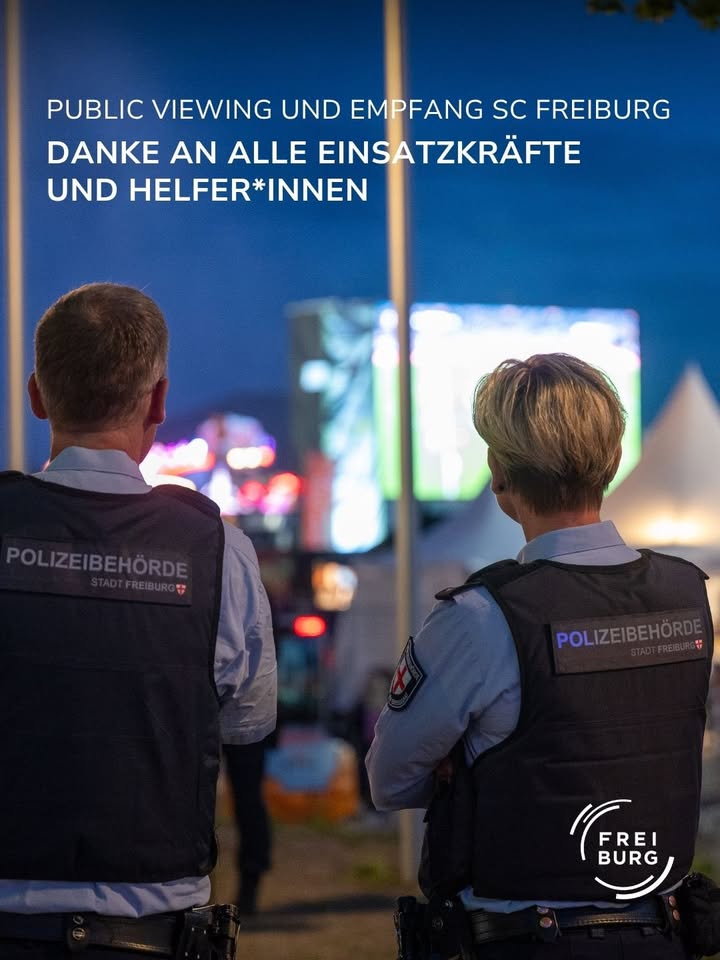 Zwei Polizist*innen der Polizeibehörde Stadt Freiburg stehen im Profil und blicken auf einen beleuchteten Bildschirm bei Nacht. Über ihnen steht der Text: „PUBLIC VIEWING UND EMPFANG SC FREIBURG – DANKE AN ALLE EINSATZKRÄFTE UND HELFER*INNEN“. Im rechten Bildrand ist das Logo „FREIBURG“ sichtbar. (KI-Alternativtext)