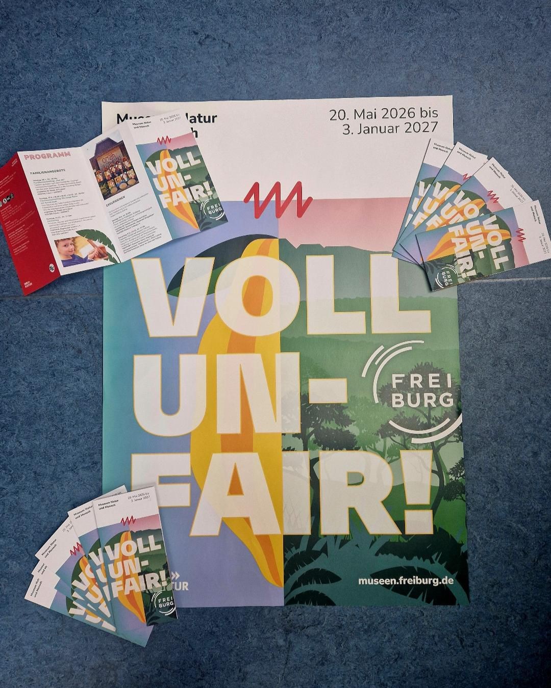 Plakat und Flyer zur Ausstellung „VOLL UN-FAIR!“ im Museum Natur und Mensch Freiburg, 20. Mai 2026 bis 3. Januar 2027, mit Programmheft und Eintrittskarten auf blauem Untergrund. (KI-Alternativtext)