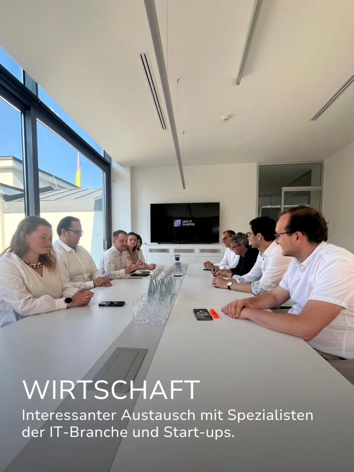 Eine Gruppe von Personen sitzt an einem langen weißen Tisch in einem hellen Raum und führt ein Gespräch. Im Hintergrund ist ein Bildschirm mit der Aufschrift „LVIV IT CLUSTER“ zu sehen. Ein Text-Overlay unten links lautet: „WIRTSCHAFT. Interessanter Austausch mit Spezialisten der IT-Branche und Start-ups.“ (KI-Alternativtext)