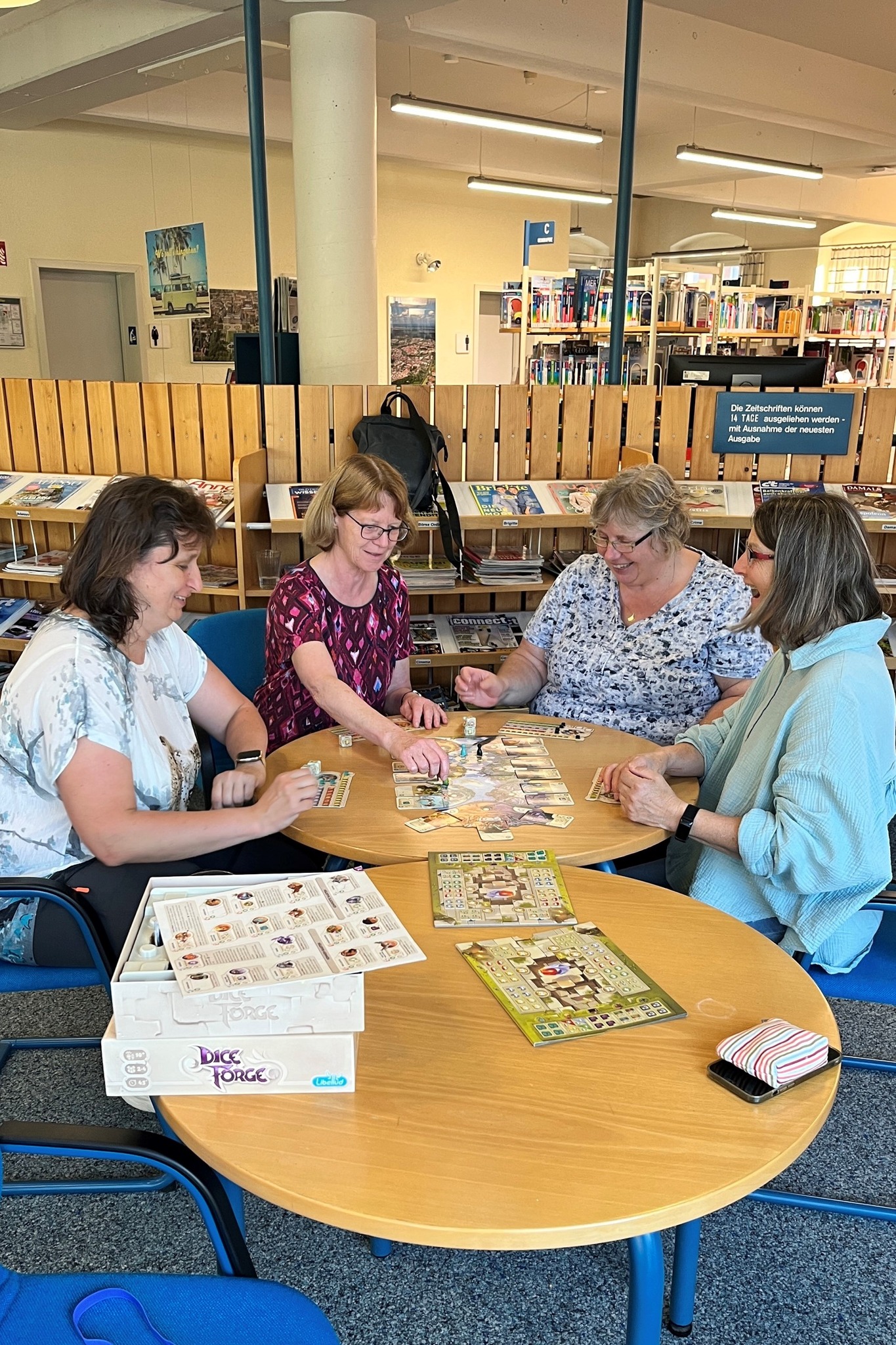 Vier Personen sitzen an einem runden Tisch in einer Bibliothek und spielen ein Brettspiel. Im Vordergrund liegt eine weitere Tischplatte mit der Spielbox "Dice Forge" und Spielplänen. Im Hintergrund sind Regale mit Zeitschriften und ein Schild mit Ausleihinformationen zu sehen. (KI-Alternativtext)