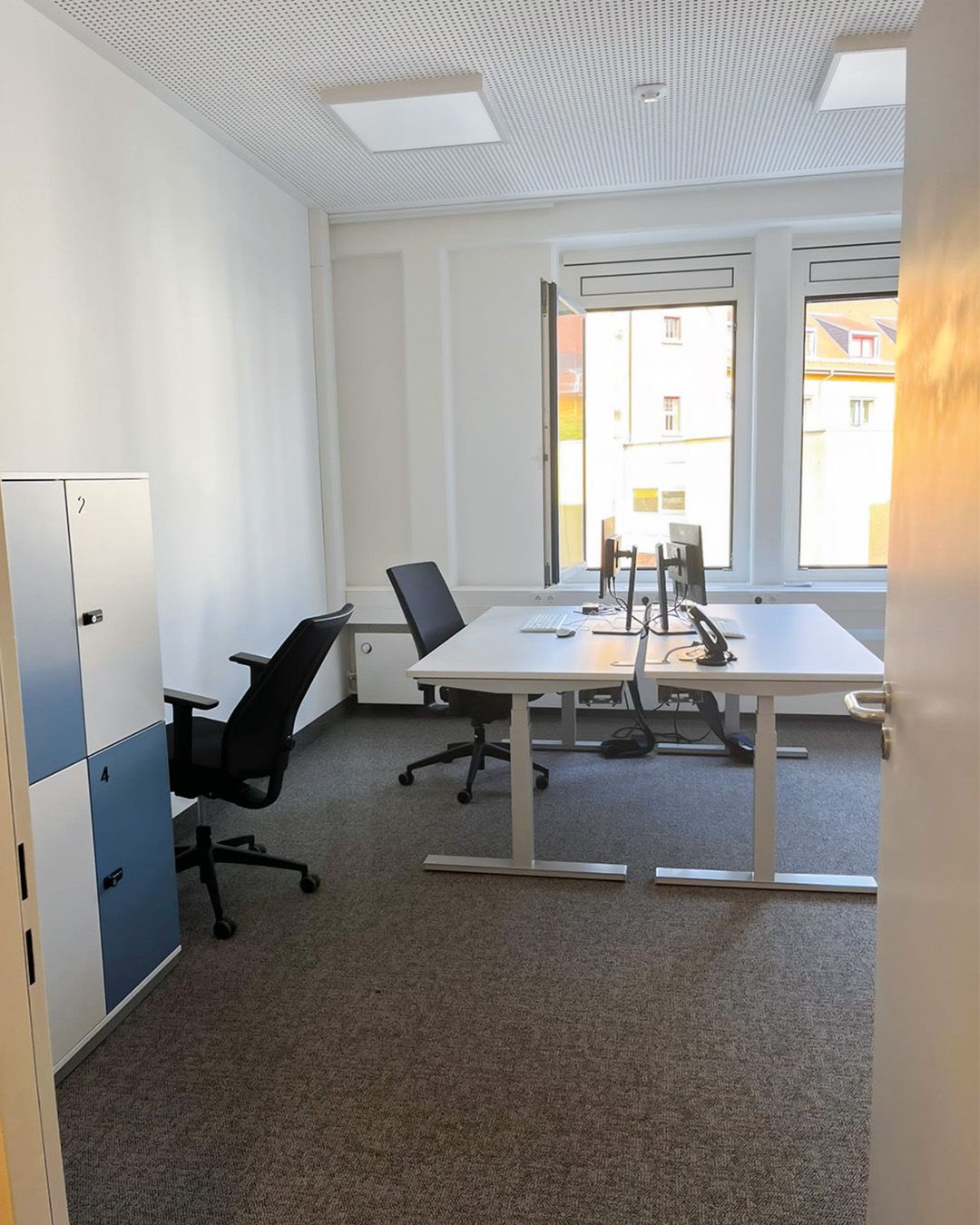 Dieser Alternativtext wurde KI-generiert: Ein heller Raum mit Schreibtischen, Stühlen und einem Aktenschrank ist zu sehen. Im Hintergrund ist durch ein Fenster ein Blick auf Gebäude im Freien möglich.