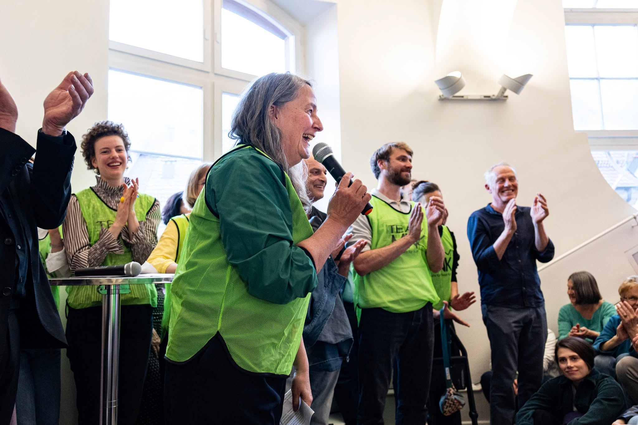Eine Person mit grauen Haaren und grüner Weste hält ein Mikrofon und lacht. Im Hintergrund klatscht eine Gruppe von Menschen, von denen viele ebenfalls grüne Westen mit der Aufschrift „TEAM“ tragen. (KI-Alternativtext)