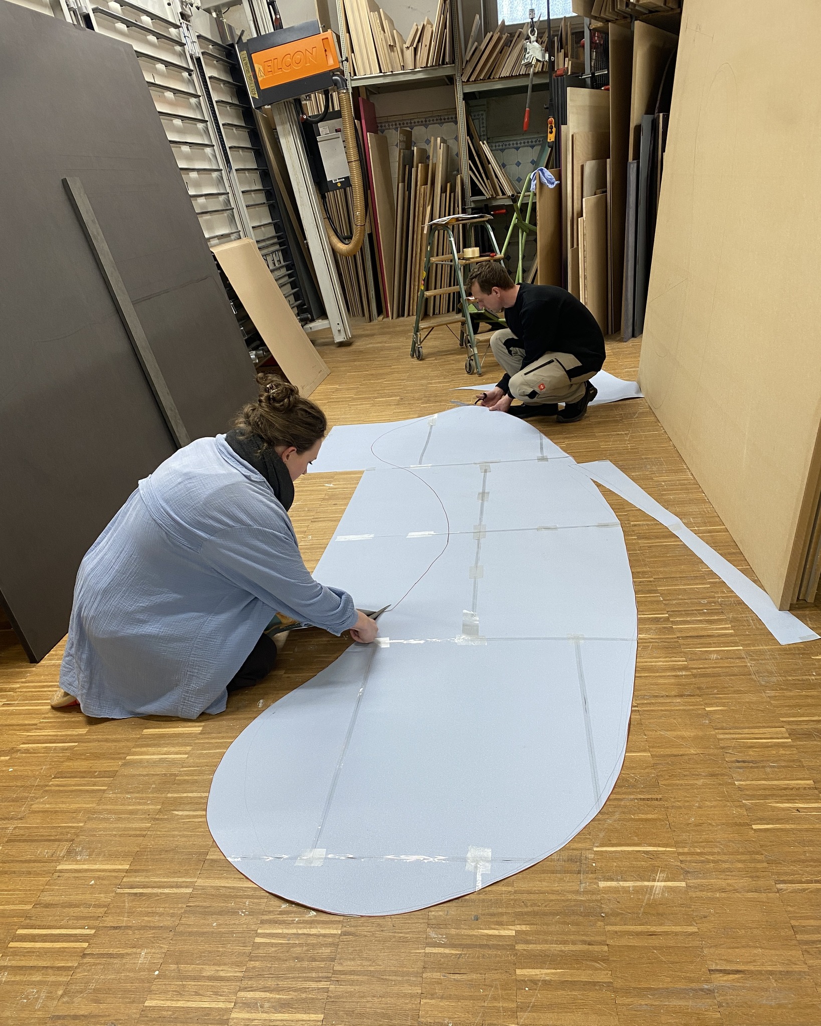 Dieser Alternativtext wurde KI-generiert: Zwei Personen arbeiten auf dem Boden eines Raumes mit verschiedenen Materialien und Werkzeugen. Im Hintergrund sind Regale mit Platten und andere Gegenstände zu sehen.