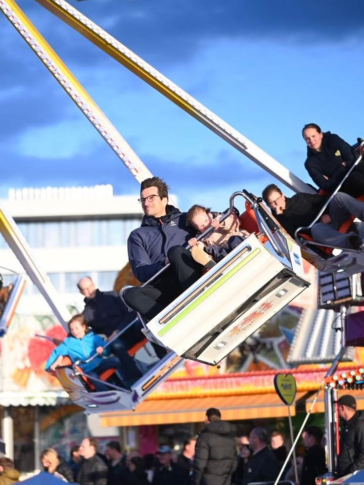 Mehrere Personen sitzen in einer schaukelnden Fahrgeschäftskabine auf einem Jahrmarkt, im Hintergrund sind weitere Besucher und Buden unter blauem Himmel zu sehen. (KI-Alternativtext)