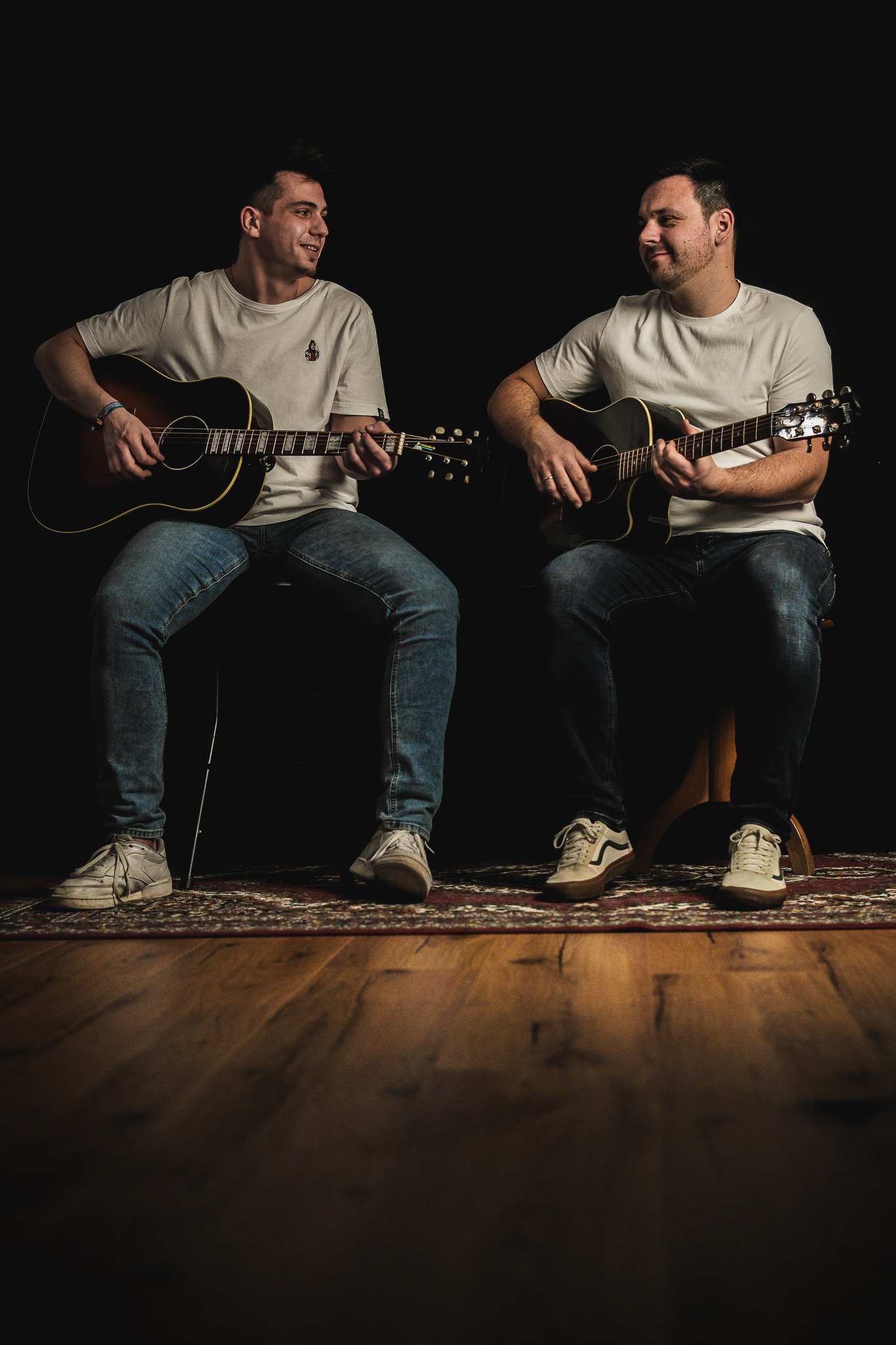 Zwei Personen in weißen T-Shirts und Jeans sitzen nebeneinander auf einem Teppich vor schwarzem Hintergrund. Beide halten akustische Gitarren, schauen sich an und lächeln. (KI-Alternativtext)