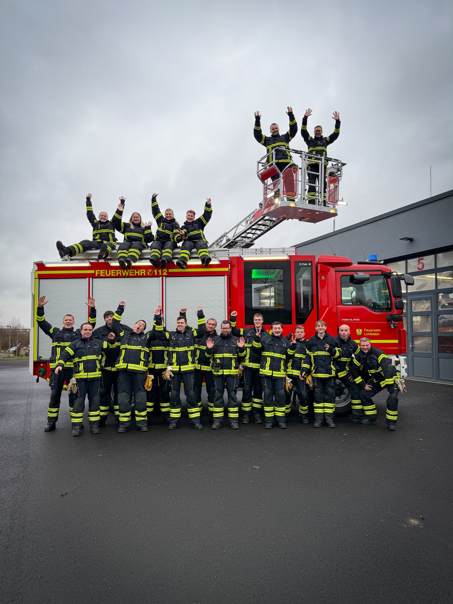 Eine Gruppe von Personen in Feuerwehruniformen posiert vor einem roten Löschfahrzeug mit der Aufschrift "FEUERWEHR 112" und "Feuerwehr Lohfelden". Mehrere Personen stehen auf dem Boden, vier sitzen auf dem Fahrzeugdach und zwei stehen in der ausgefahrenen Drehleiter, wobei viele in die Kamera winken. (KI-Alternativtext)