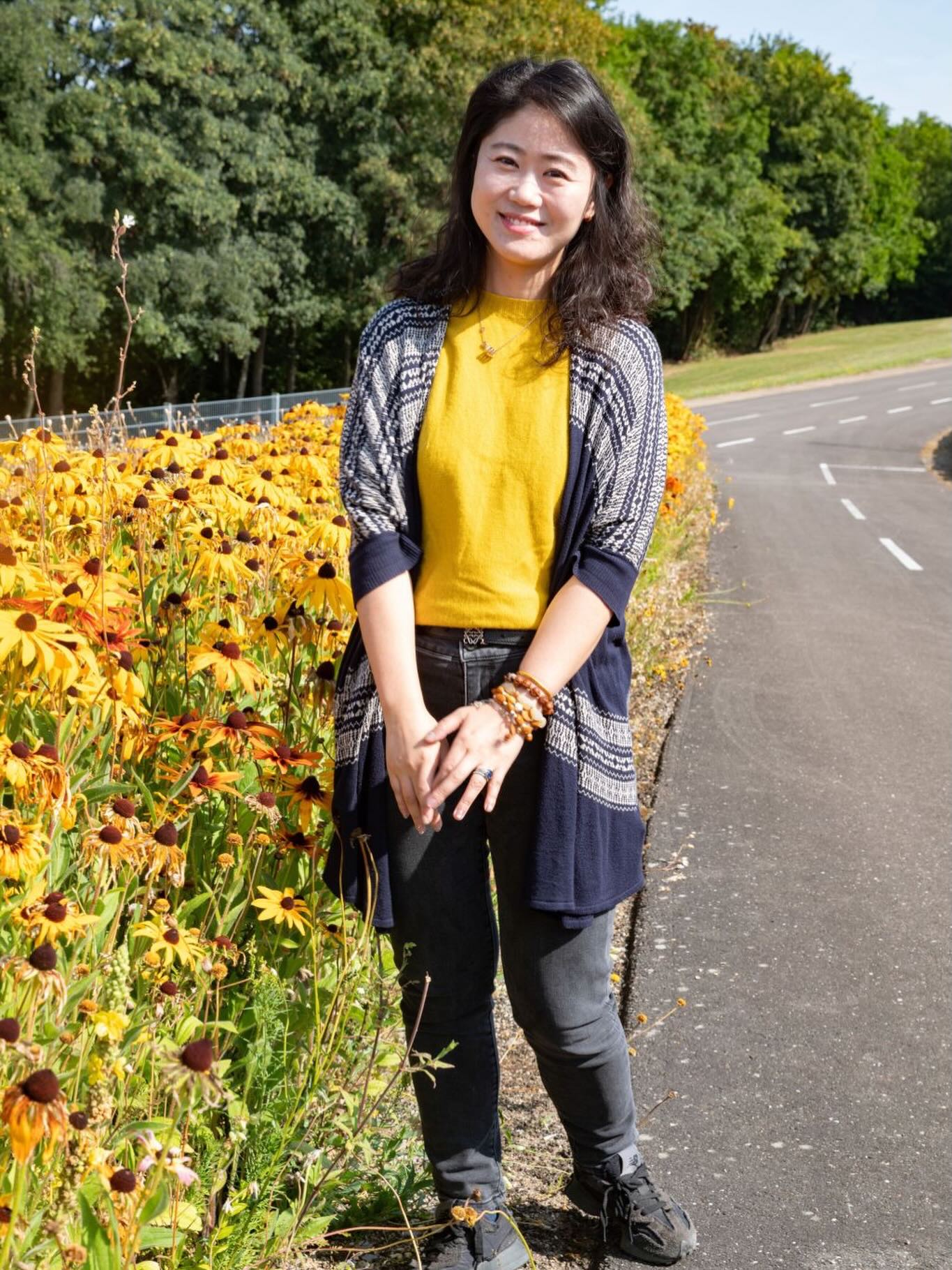 Eine Person mit dunklen, schulterlangen Haaren steht lächelnd im Freien neben einem Beet mit gelben Blumen. Die Person trägt ein gelbes Oberteil, eine dunkelblaue Strickjacke mit Muster und eine dunkle Hose. Rechts verläuft ein asphaltierter Weg, im Hintergrund sind Bäume zu sehen. (KI-Alternativtext)