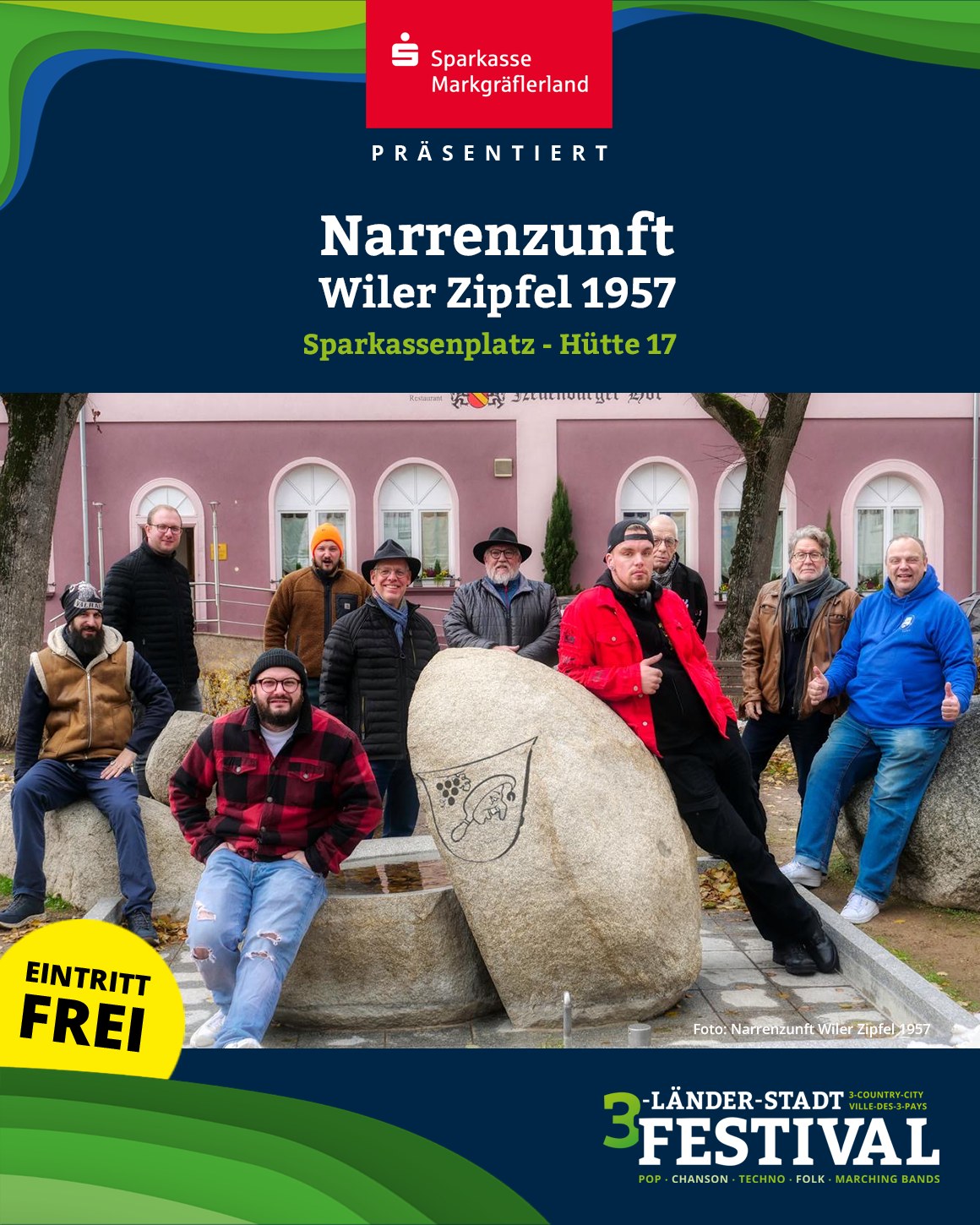 Ein Gruppenfoto von mehreren Personen im Freien vor einem Gebäude und einem Steindenkmal. Im Vordergrund steht ein gelber Kreis mit der Aufschrift "Eintritt Frei".