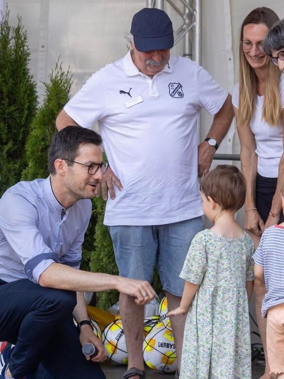 Eine Person in einem hellblauen Hemd hockt und hält ein Mikrofon, während sie mit einem kleinen Kind in einem Blumenkleid interagiert. Im Hintergrund stehen weitere Personen, darunter eine in einem weißen Poloshirt und einer Kopfbedeckung. Am Boden liegen zwei gelbe Bälle mit der Aufschrift „DERBY'S“. (KI-Alternativtext)