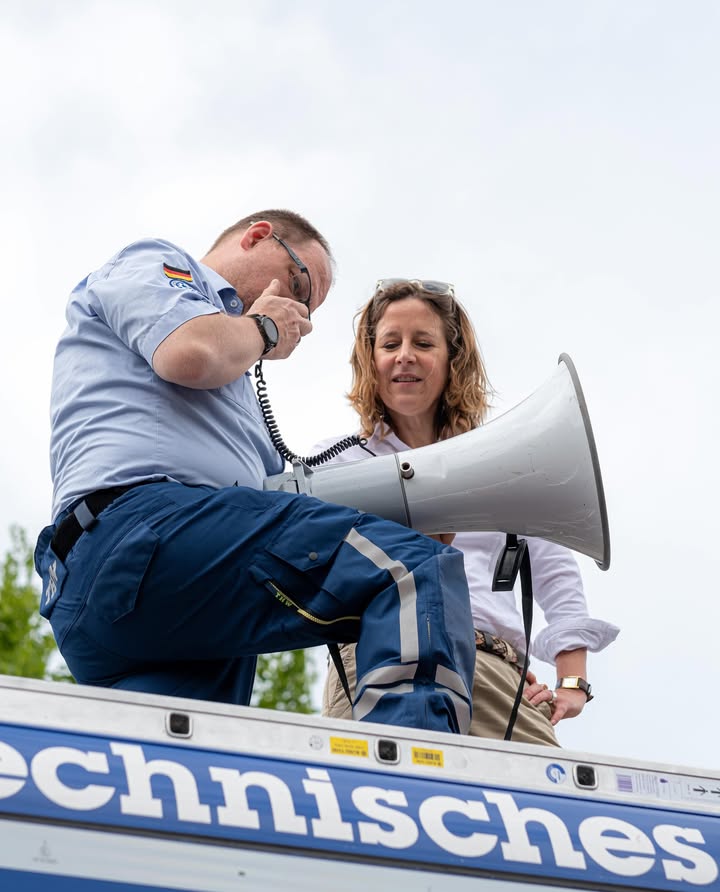 Dieser Alternativtext wurde KI-generiert: Zwei Personen in Uniformen befinden sich auf einem Fahrzeugdach. Eine Person spricht in ein Megafon, während die andere am Telefon zu sein scheint.