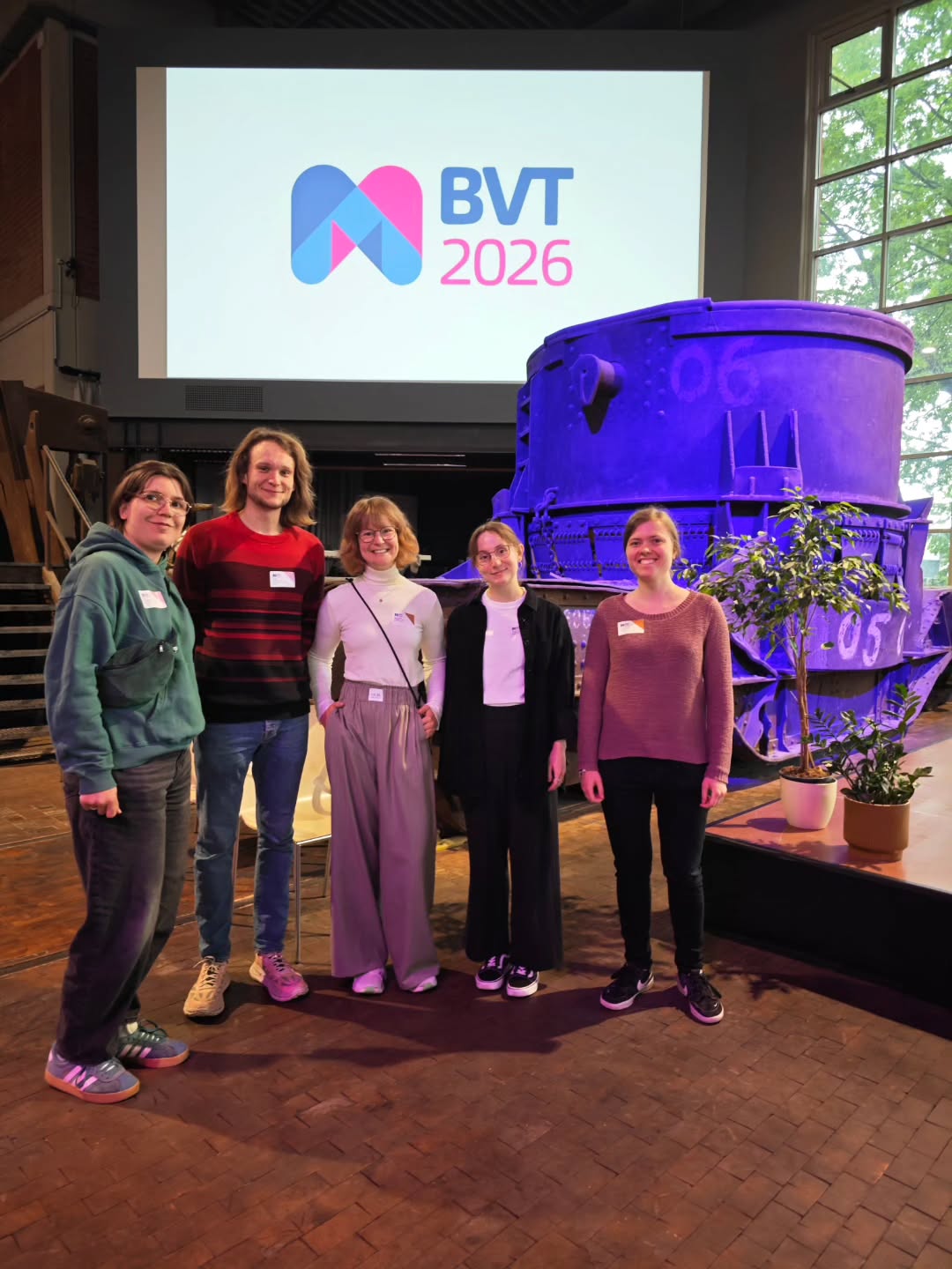 Fünf Personen stehen vor einer Projektionsleinwand mit dem Logo „BVT 2026“ und einer großen blauen Maschine. Alle tragen Namensschilder und blicken in die Kamera. Im Hintergrund sind Fenster und Pflanzen sichtbar. (KI-Alternativtext)
