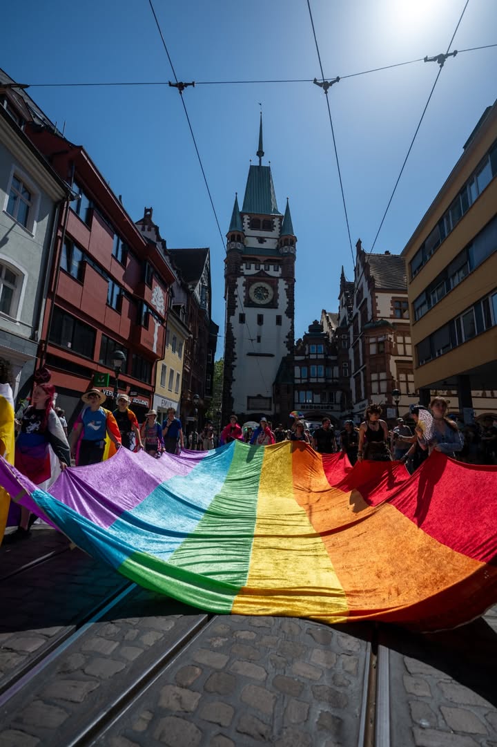 Menschen halten eine große Regenbogenfahne auf einer Straße mit Straßenbahnschienen, im Hintergrund ein markanter Uhrturm und historische Gebäude unter klarem Himmel. (KI-Alternativtext)