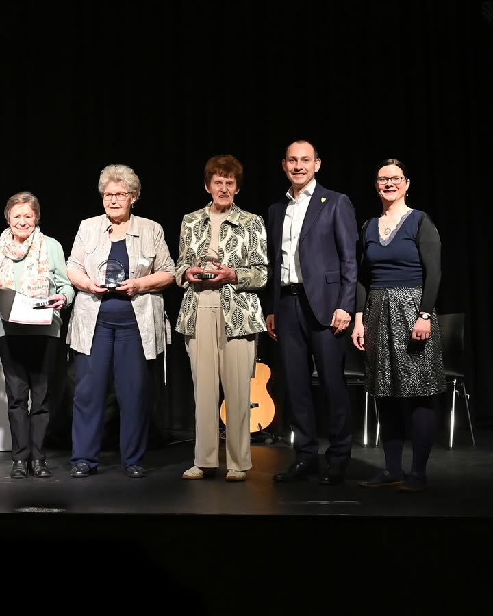Dieser Alternativtext wurde KI-generiert: Eine Gruppe von fünf Personen steht auf einer Bühne. Einige halten Auszeichnungen in den Händen und lächeln in die Kamera.