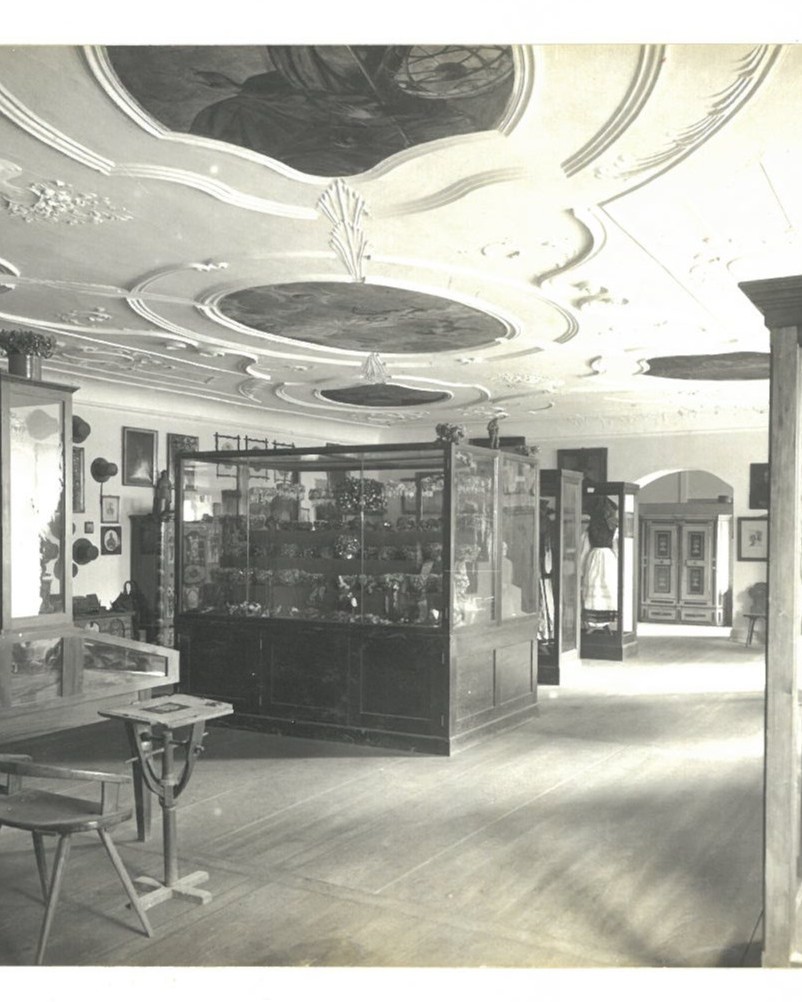 Dieser Alternativtext wurde KI-generiert: Schwarz-weißes Bild eines Innenraumes mit kunstvollen Stuckverzierungen an der Decke. Im Raum befinden sich Vitrinen und Bilder an der Wand.