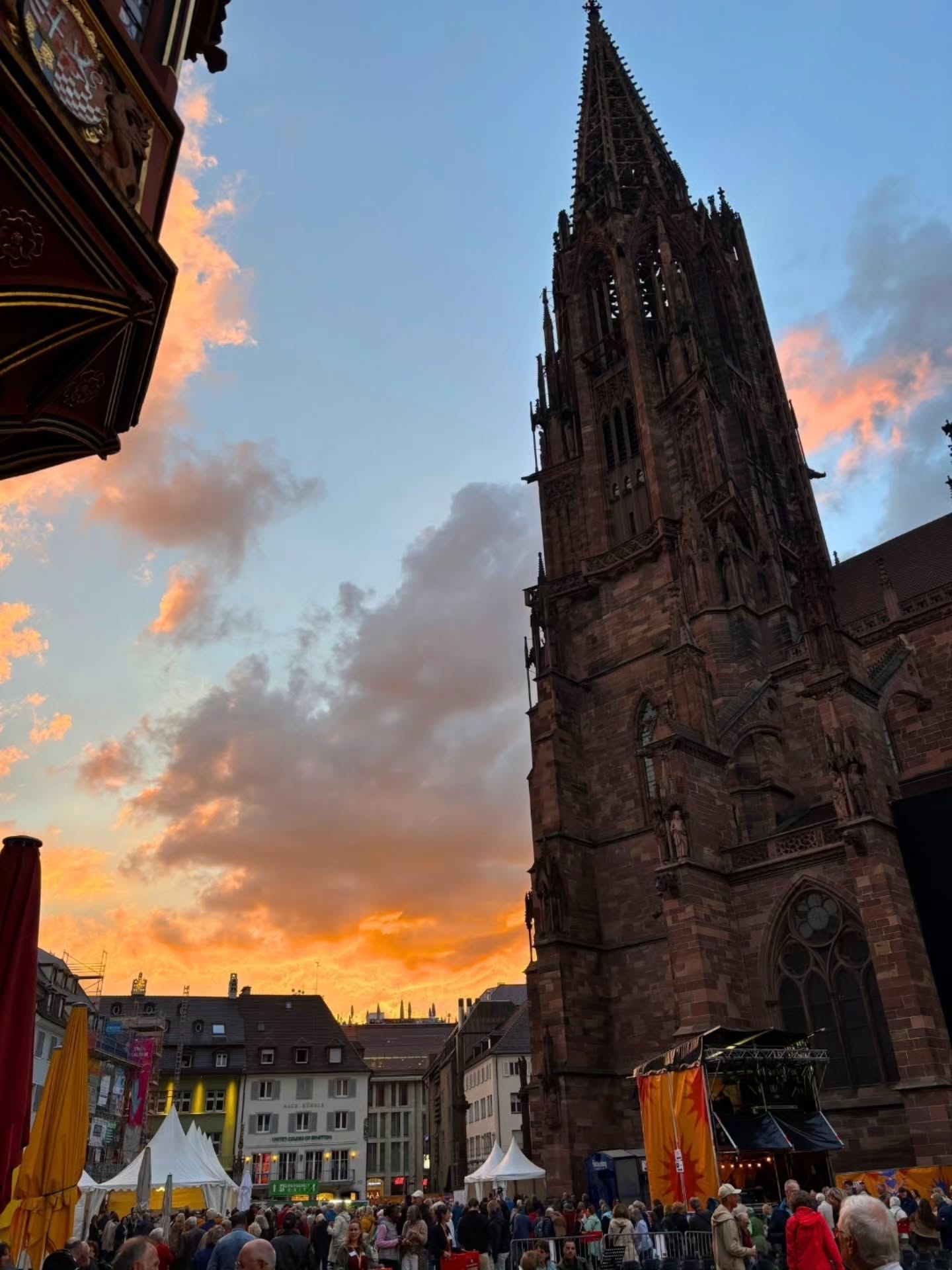 Hoher gotischer Kirchturm vor einem orangefarbenen Abendhimmel, im Vordergrund Menschenmenge auf einem belebten Platz mit Zelten und Bühne. (KI-Alternativtext)