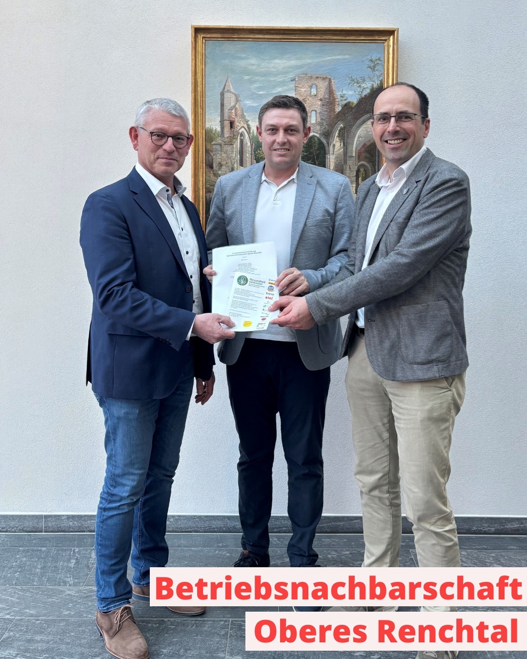 Dieser Alternativtext wurde KI-generiert: Drei Personen in formeller Kleidung stehen nebeneinander und halten Dokumente. Im Hintergrund ist ein Gemälde mit einer Kirche und einer Burgruine zu sehen.