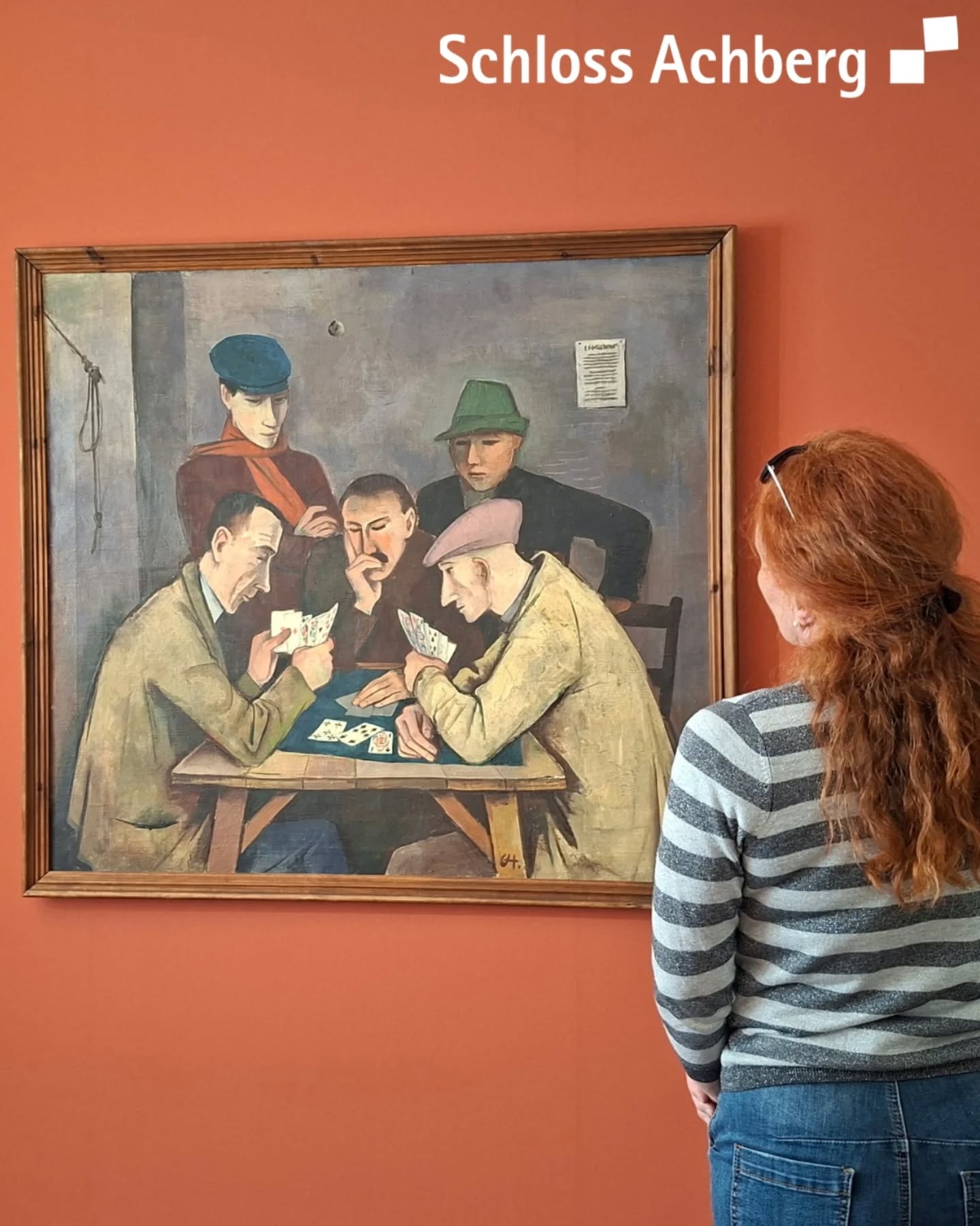 Dieser Alternativtext wurde KI-generiert: Ein gerahmtes Gemälde zeigt eine Gruppe von Personen, die an einem Tisch sitzen und spielen. Eine Person steht seitlich davon und betrachtet das Bild.