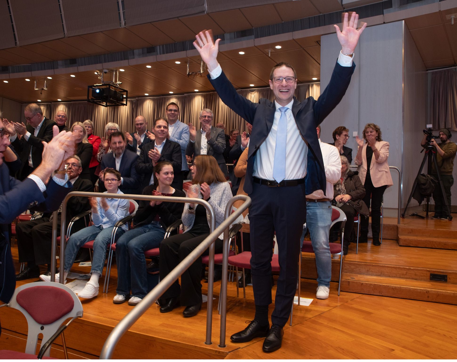 Dieser Alternativtext wurde KI-generiert: Eine Person steht auf einer Bühne mit erhobenen Armen, umgeben von einer Menschenmenge, die applaudiert. Im Hintergrund befindet sich ein Innenraum mit Stühlen und einer Treppe.