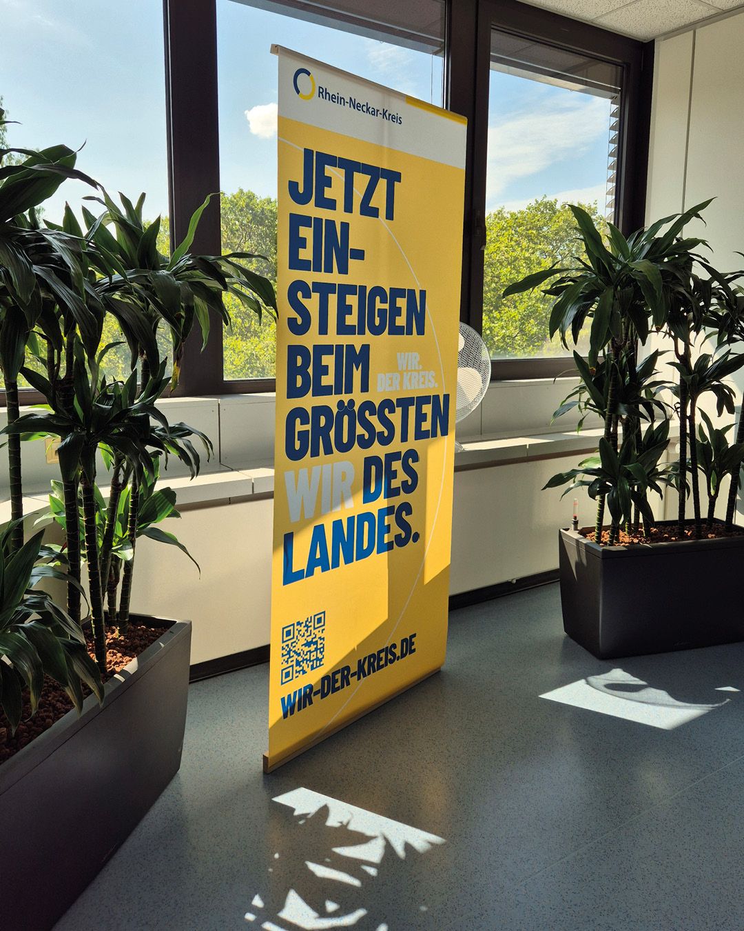 Ein gelber Roll-up steht zwischen zwei großen Topfpflanzen vor einem Fenster. Der Text lautet: „Rhein-Neckar-Kreis“, „JETZT EINSTEIGEN BEIM GRÖSSTEN DES LANDES.“ sowie „WIR. DER KREIS.“. Unten befindet sich ein QR-Code und die Adresse „WIR-DER-KREIS.DE“. (KI-Alternativtext)