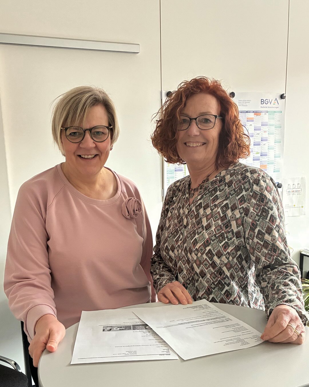 Dieser Alternativtext wurde KI-generiert: Zwei lächelnde Personen stehen nebeneinander und halten Dokumente in der Hand. Im Hintergrund ist ein Schreibtisch mit einem Kalender und weiteren Papieren zu sehen.