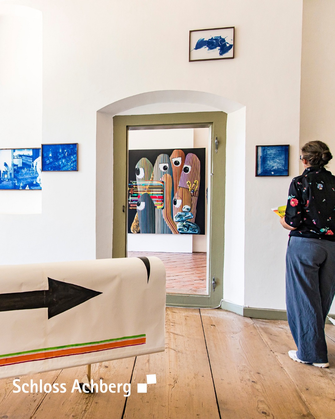 Dieser Alternativtext wurde KI-generiert: Ein Innenraum mit einer Türöffnung und mehreren Bildern an den Wänden. Der Raum hat einen Holzboden und ein auffälliges Bodendesign mit einem Pfeil.