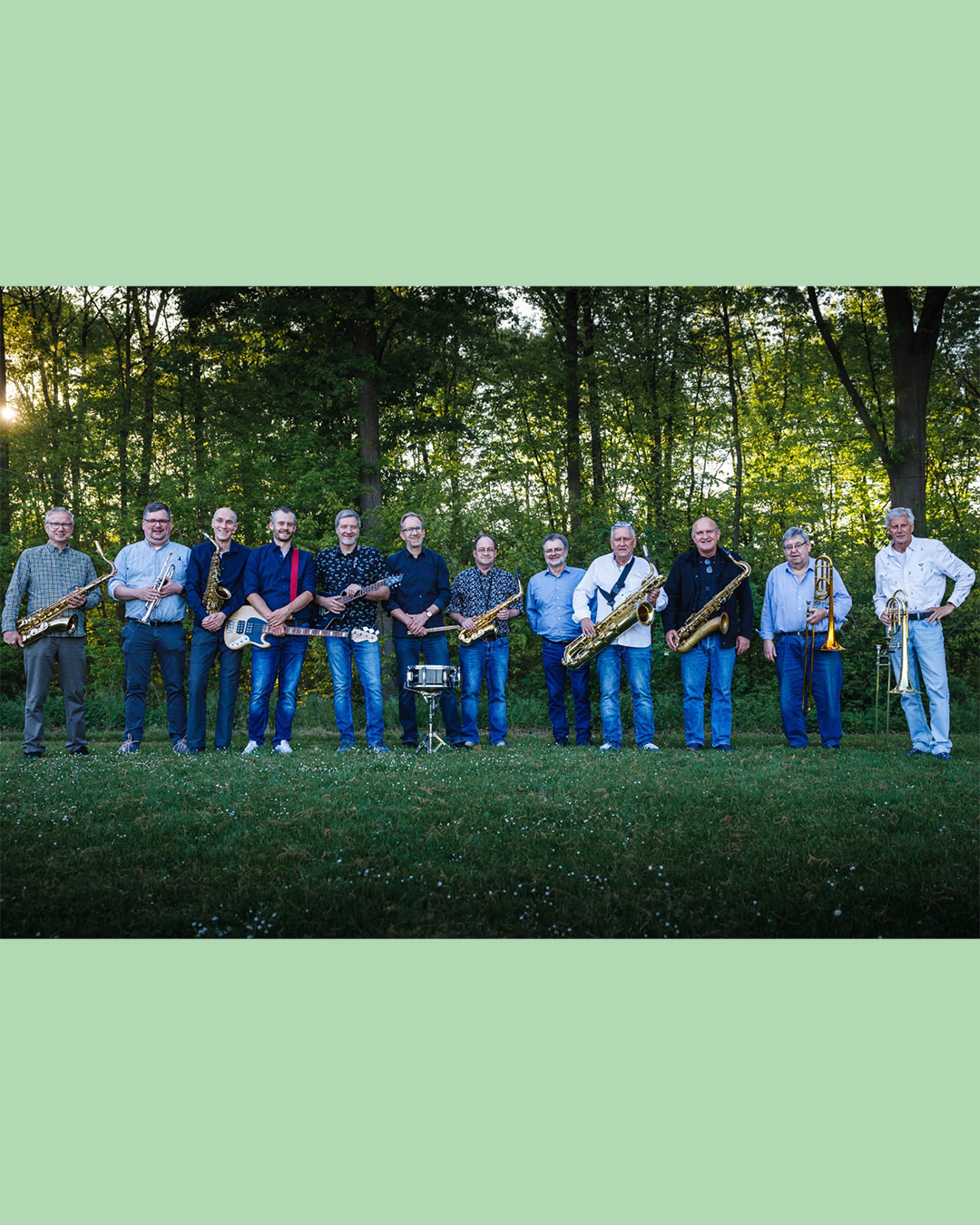 Zwölf Personen stehen in einer Reihe auf einer Wiese vor einem Waldrand. Sie halten verschiedene Instrumente, darunter Saxophone, Trompeten, eine Posaune und E-Gitarren. In der Mitte steht ein kleines Schlaginstrument auf einem Ständer. (KI-Alternativtext)