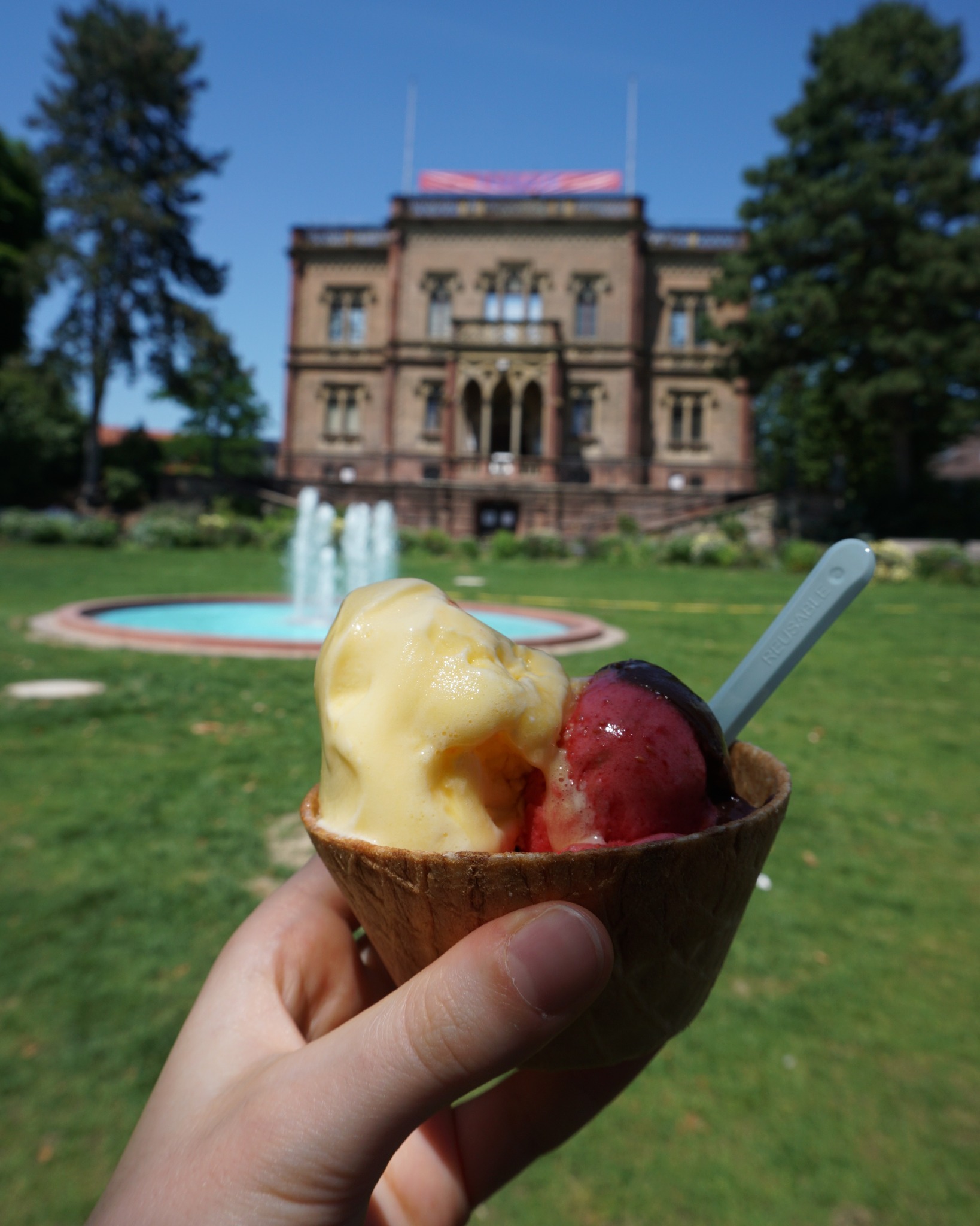 Eine Hand hält eine Waffelschale mit zwei Kugeln Eiscreme – einer gelb, eine rot – und einem blauen Löffel. Im Hintergrund ist ein großes Gebäude mit Brunnen und grünem Rasen unter blauem Himmel zu sehen. (KI-Alternativtext)