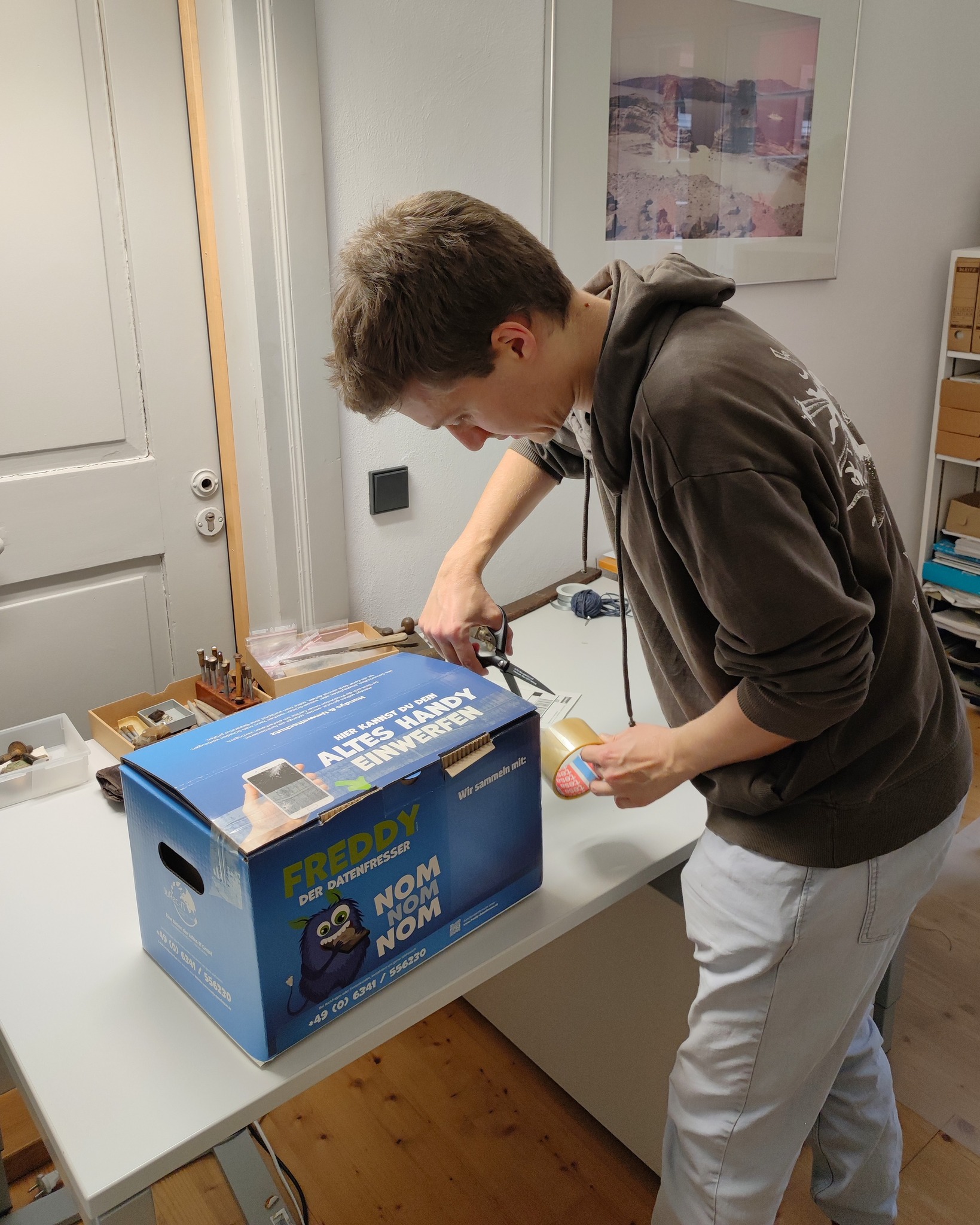 Dieser Alternativtext wurde KI-generiert: Eine Person steht in einem Raum und betrachtet einen geöffneten Karton auf einer weißen Oberfläche. Der Raum enthält Möbel und eine Tür.