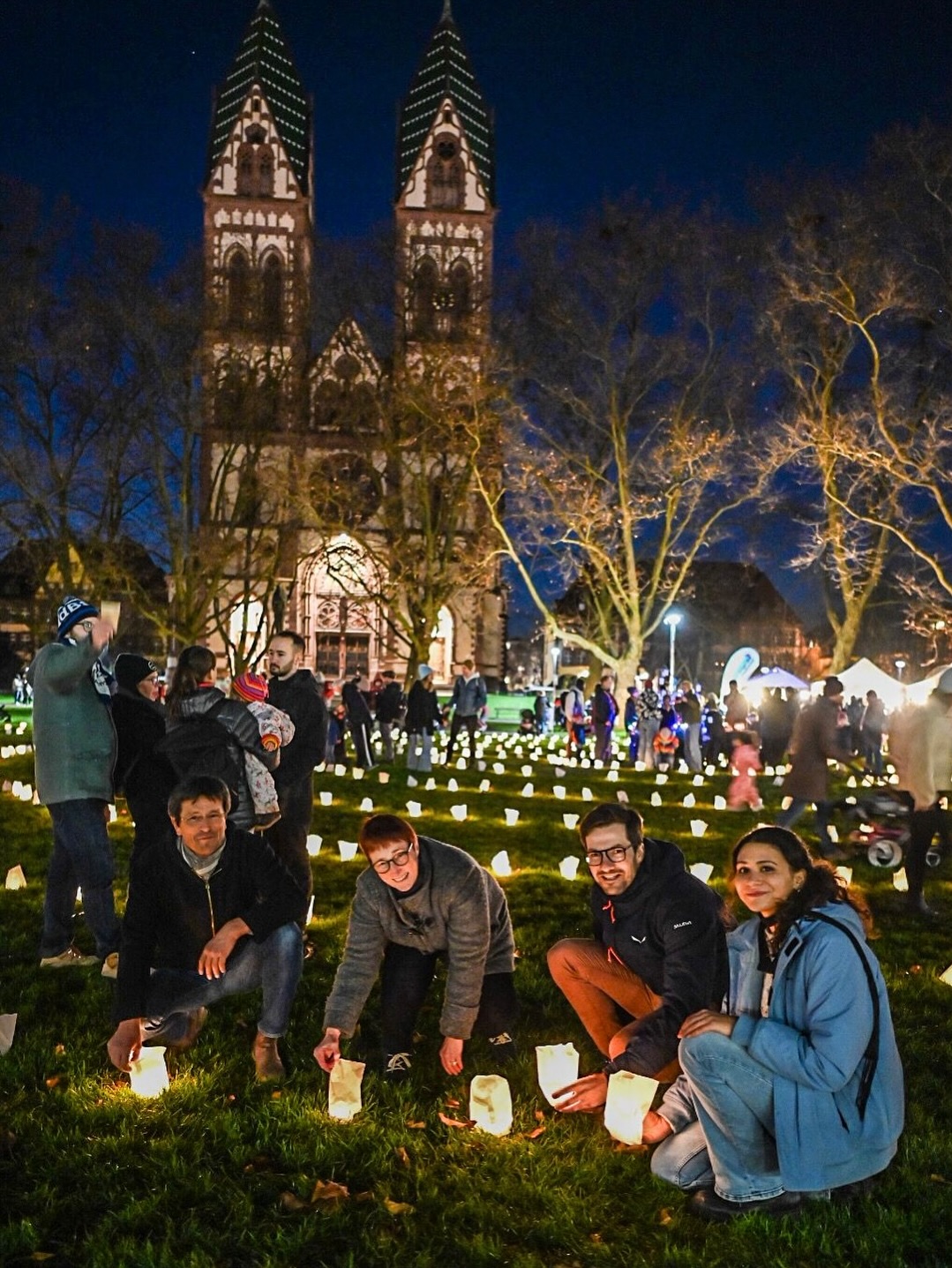 Dieser Alternativtext wurde KI-generiert: Ein beleuchtetes Bauwerk im Hintergrund wird von einer Menschengruppe in einem Park mit vielen kleinen Lichtern umgeben. Es scheint eine Veranstaltung im Freien zu sein.