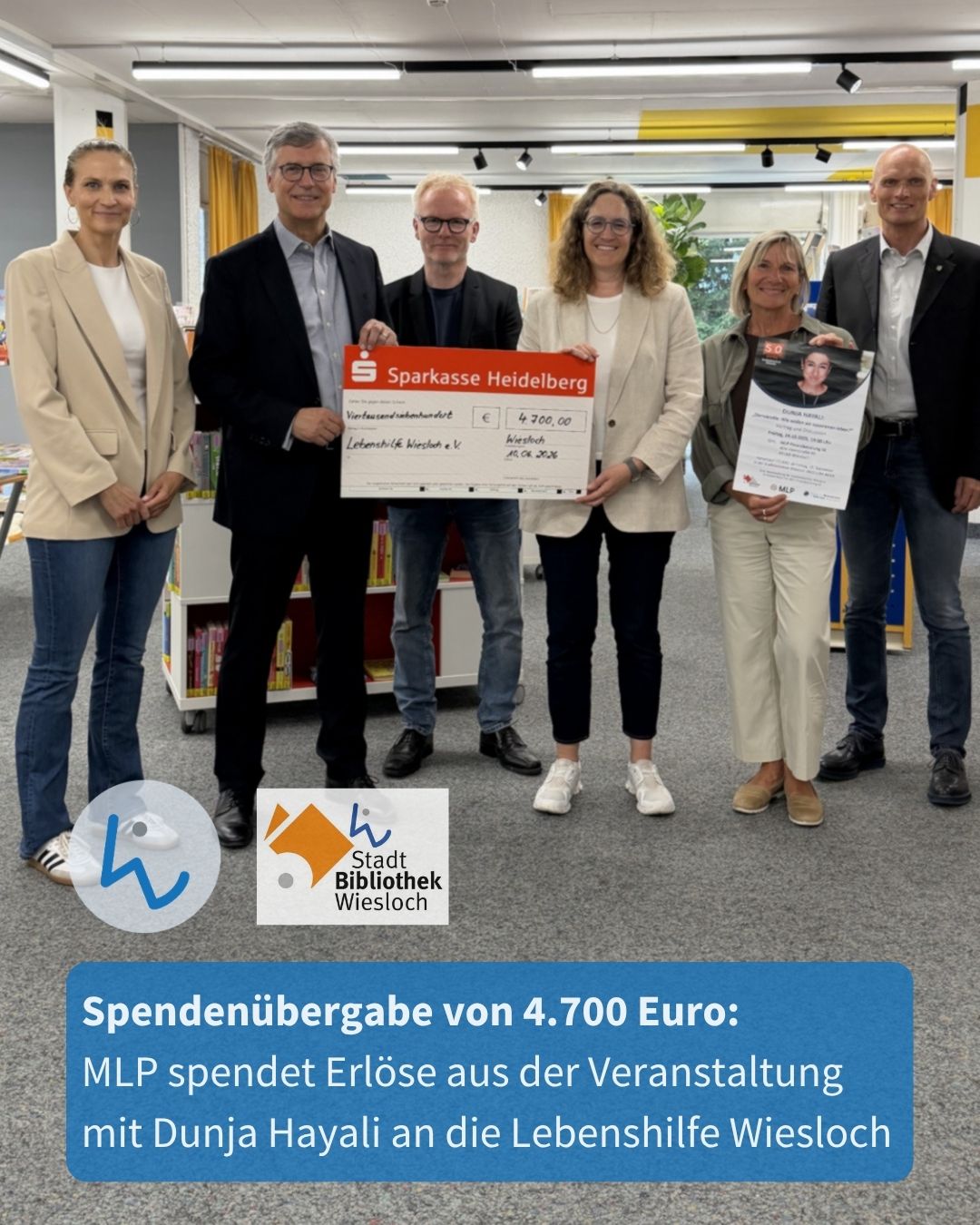 Sechs Personen stehen in einem Raum mit Bücherregalen und halten einen großen Spendenscheck der Sparkasse Heidelberg über 4.700 Euro an die Lebenshilfe Wiesloch sowie ein Plakat mit einem Porträt. Unten sind Logos der Stadt Bibliothek Wiesloch und ein Textfeld mit der Spendendetails. (KI-Alternativtext)