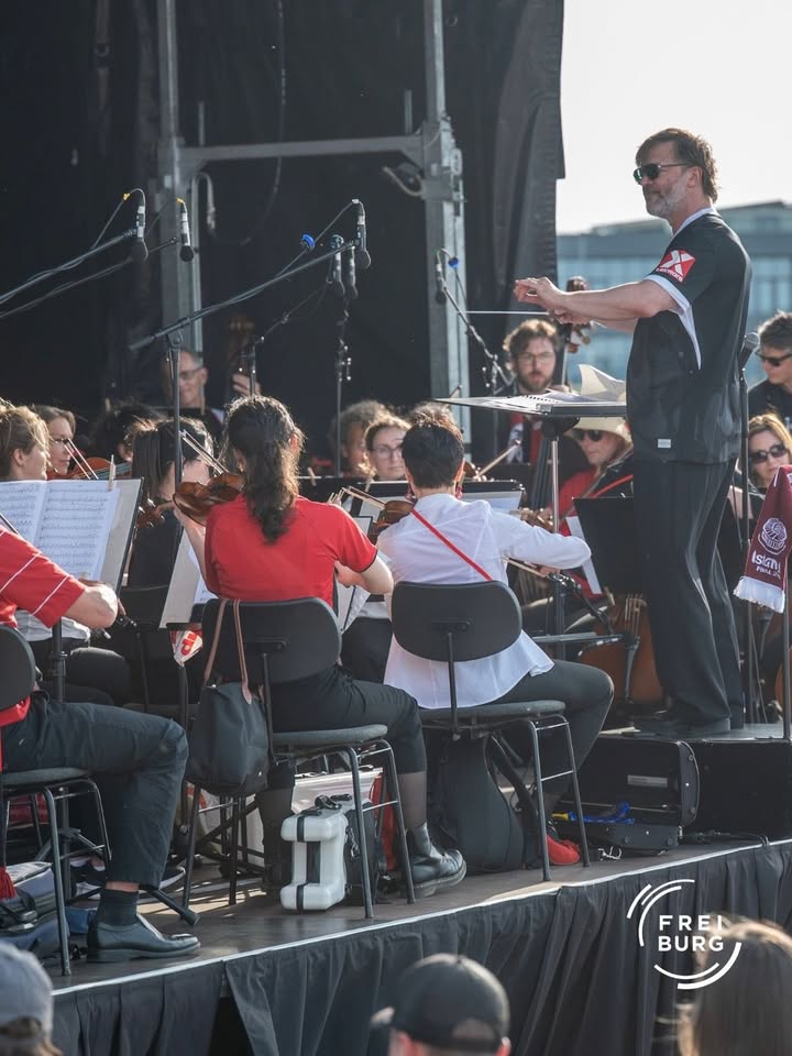 Dirigent mit Sonnenbrille und Stab leitet ein Orchester auf einer Bühne, Musiker spielen Streichinstrumente, einige tragen rote Oberteile, im Hintergrund Mikrofone und Bühnentechnik. (KI-Alternativtext)