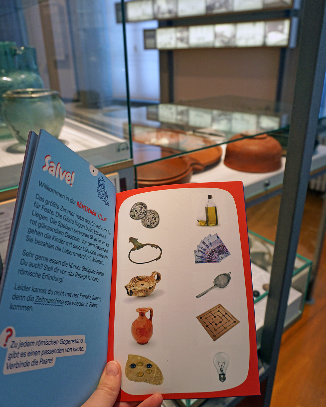 Dieser Alternativtext wurde KI-generiert: Ein Schild mit Text und Illustrationen steht in einer Vitrine. Es zeigt Szenen aus der römischen Zeit und Informationen zu einem Spiel.