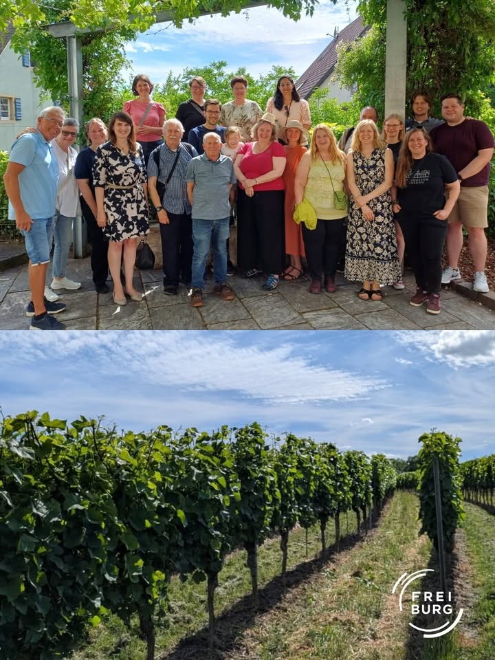 Gruppe von Menschen posiert im Freien unter einem Pergola, dahinter ein Weingarten mit Reihen von Rebstöcken unter blauem Himmel; rechts unten Logo „FREIBURG“. (KI-Alternativtext)