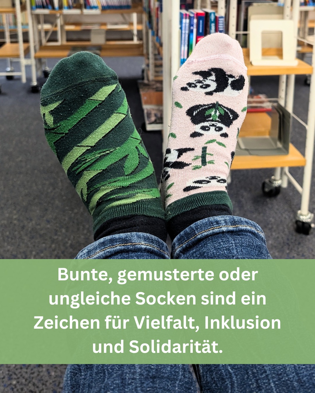 Bild von zwei unterschiedlich gemusterten Socken an Füßen, die auf einer Jeanshose liegen.  Der Hintergrund zeigt eine Bücherei.