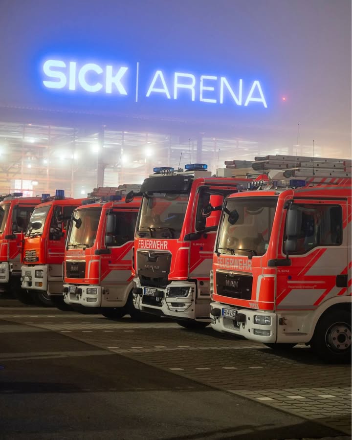 Dieser Alternativtext wurde KI-generiert: Mehrere rote Fahrzeuge mit Blaulicht stehen vor einem Gebäude mit einer hellen blauen Leuchtreklame. Das Bild ist bei Nacht aufgenommen und wirkt durch den Nebel etwas verschwommen.