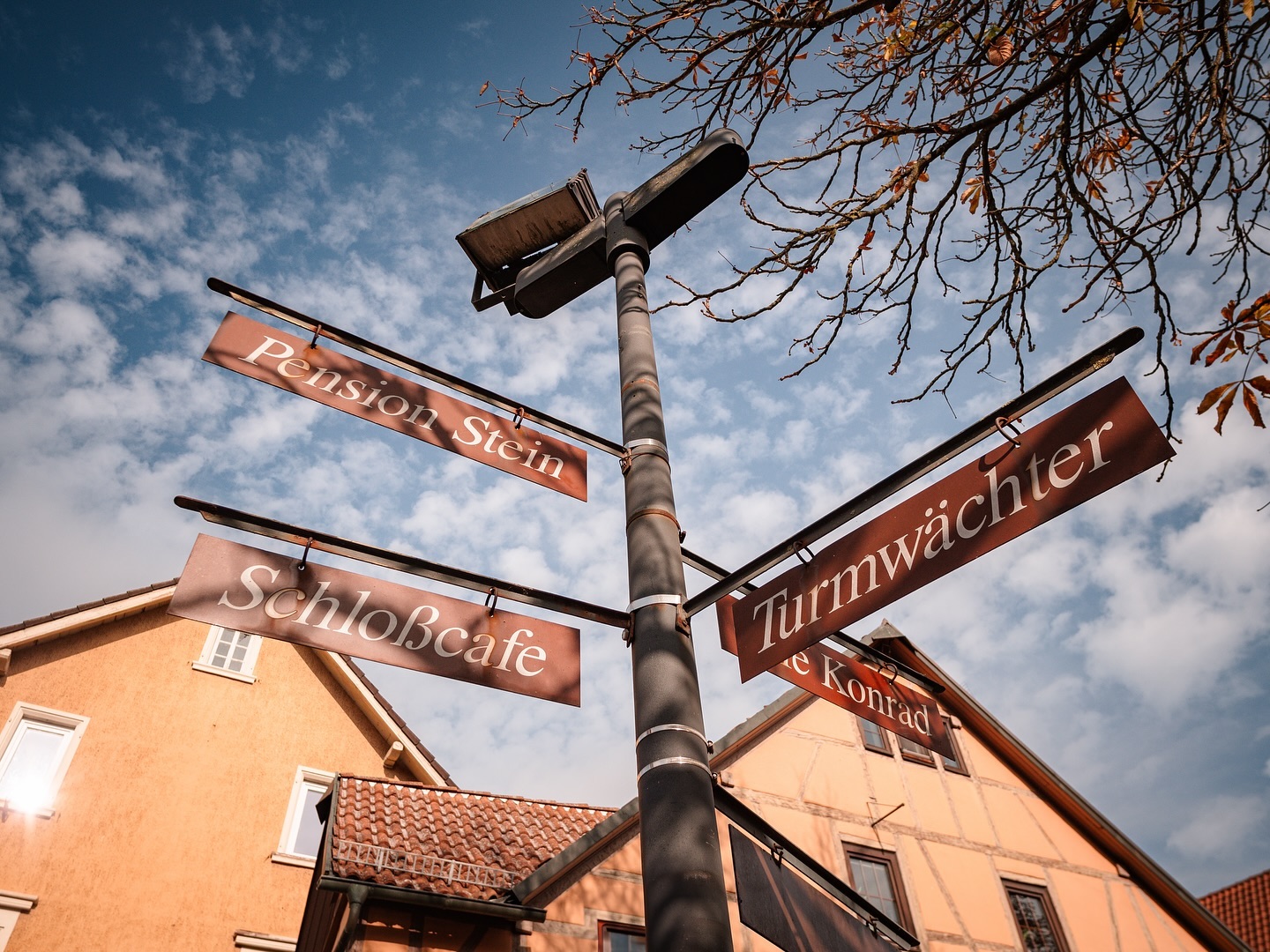 Wegweiser mit mehreren Schildern an einem Mast vor Häusern. Der Himmel ist leicht bewölkt.