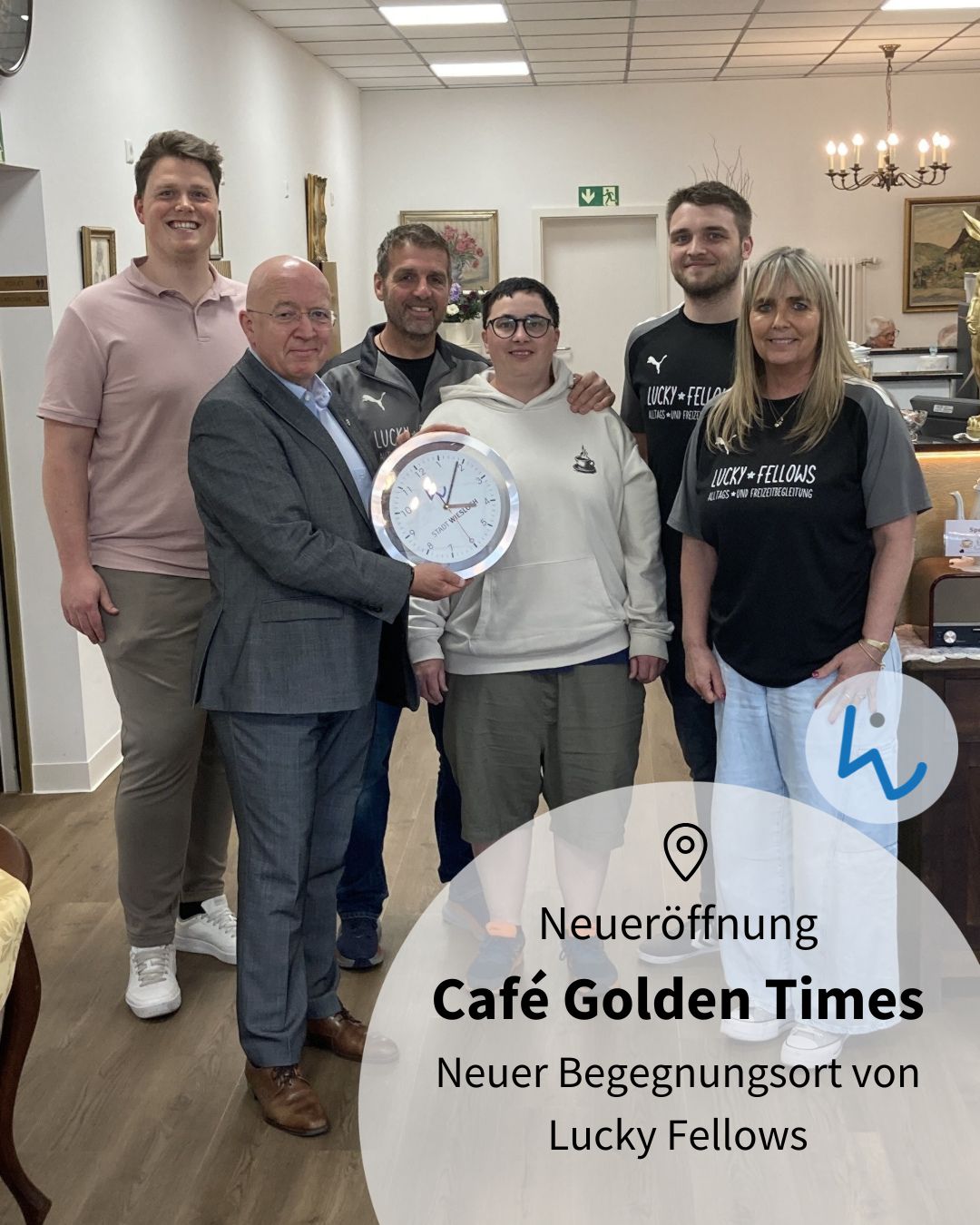 Gruppe von sechs Personen in einem Café, eine hält eine große Uhr mit der Aufschrift „Stadt Wiesbaden“. Im Vordergrund Text: „Neueröffnung Café Golden Times, Neuer Begegnungsort von Lucky Fellows“. (KI-Alternativtext)