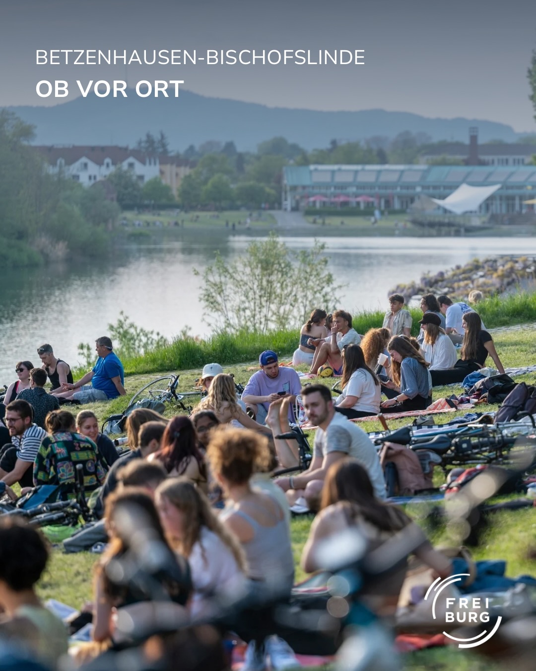 Dieser Alternativtext wurde KI-generiert: Eine größere Gruppe von Menschen sitzt und entspannt sich auf einer Wiese am Ufer eines Gewässers. Im Hintergrund ist ein Gebäude mit einem roten Giebel zu sehen.