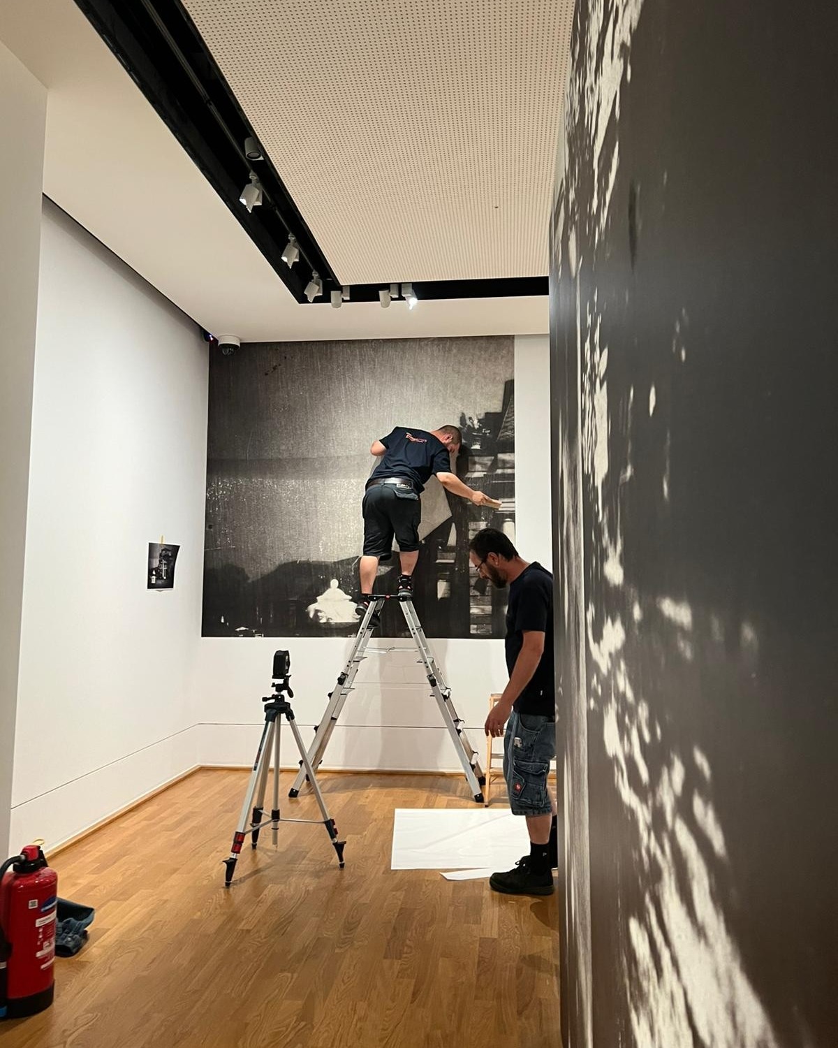 Zwei Personen montieren in einem Ausstellungsraum ein großes Schwarz-Weiß-Foto an der Wand; eine steht auf einer Leiter, die andere beobachtet von unten. Im Raum stehen ein Stativ, ein Feuerlöscher und ein kleines Bild an der Wand. (KI-Alternativtext)