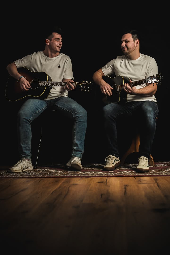 Zwei Personen in hellen T-Shirts und Jeans sitzen nebeneinander auf einem Teppich vor einem schwarzen Hintergrund. Beide halten Akustikgitarren und schauen sich lächelnd an. (KI-Alternativtext)