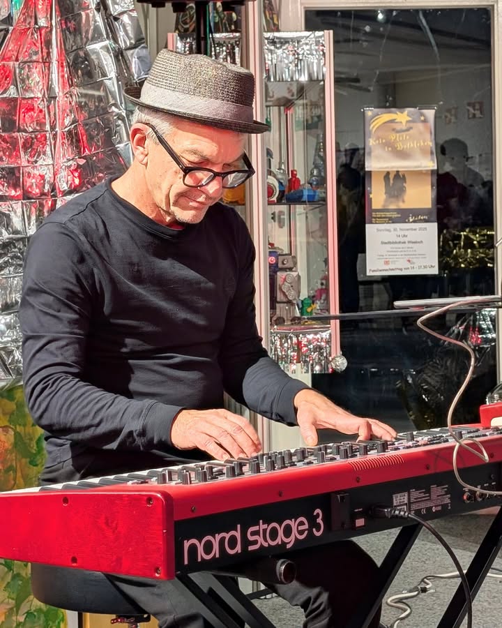 Dieser Alternativtext wurde KI-generiert: Eine Person spielt auf einem roten Keyboard im Freien. Im Hintergrund ist eine beleuchtete Verkaufsvitrinen und ein Ankündigungsplakat zu sehen.