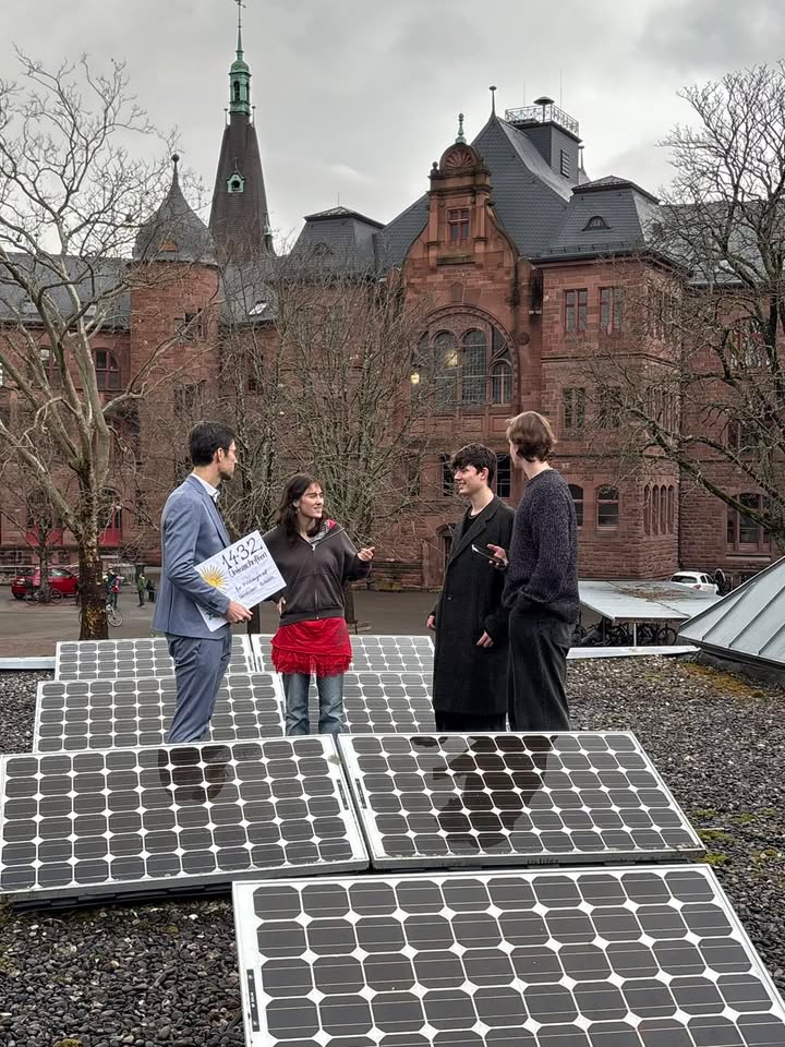 Dieser Alternativtext wurde KI-generiert: Ein Gebäude mit roten Backsteinwänden bildet den Hintergrund für eine Gruppe von Personen, die vor Solarmodulen stehen und sich unterhalten. Im Vordergrund sind mehrere Solarpanele zu sehen.