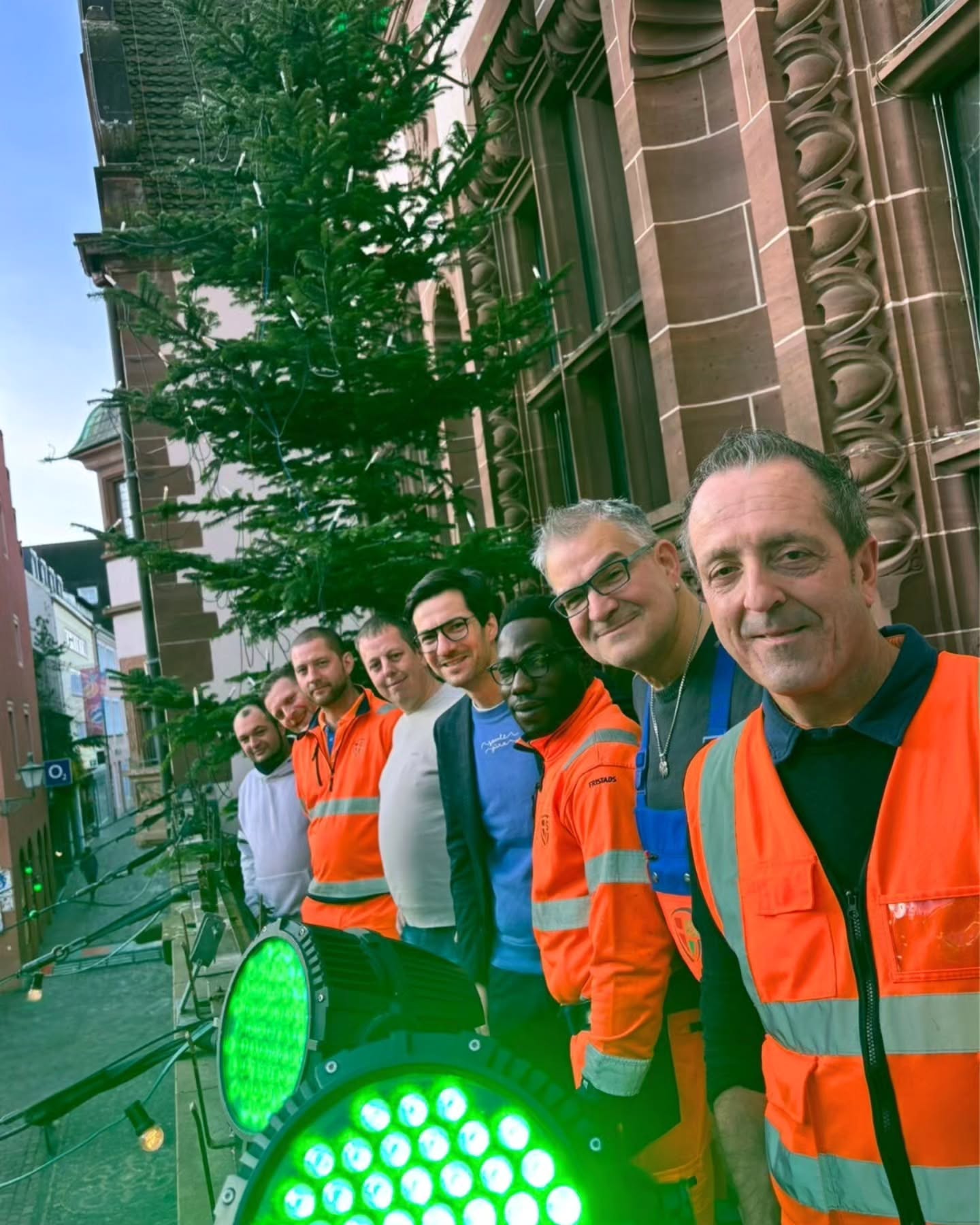Dieser Alternativtext wurde KI-generiert: Ein Weihnachtsbaum steht vor einem Gebäude. Mehrere Personen in Warnwesten stehen davor und lächeln in die Kamera.