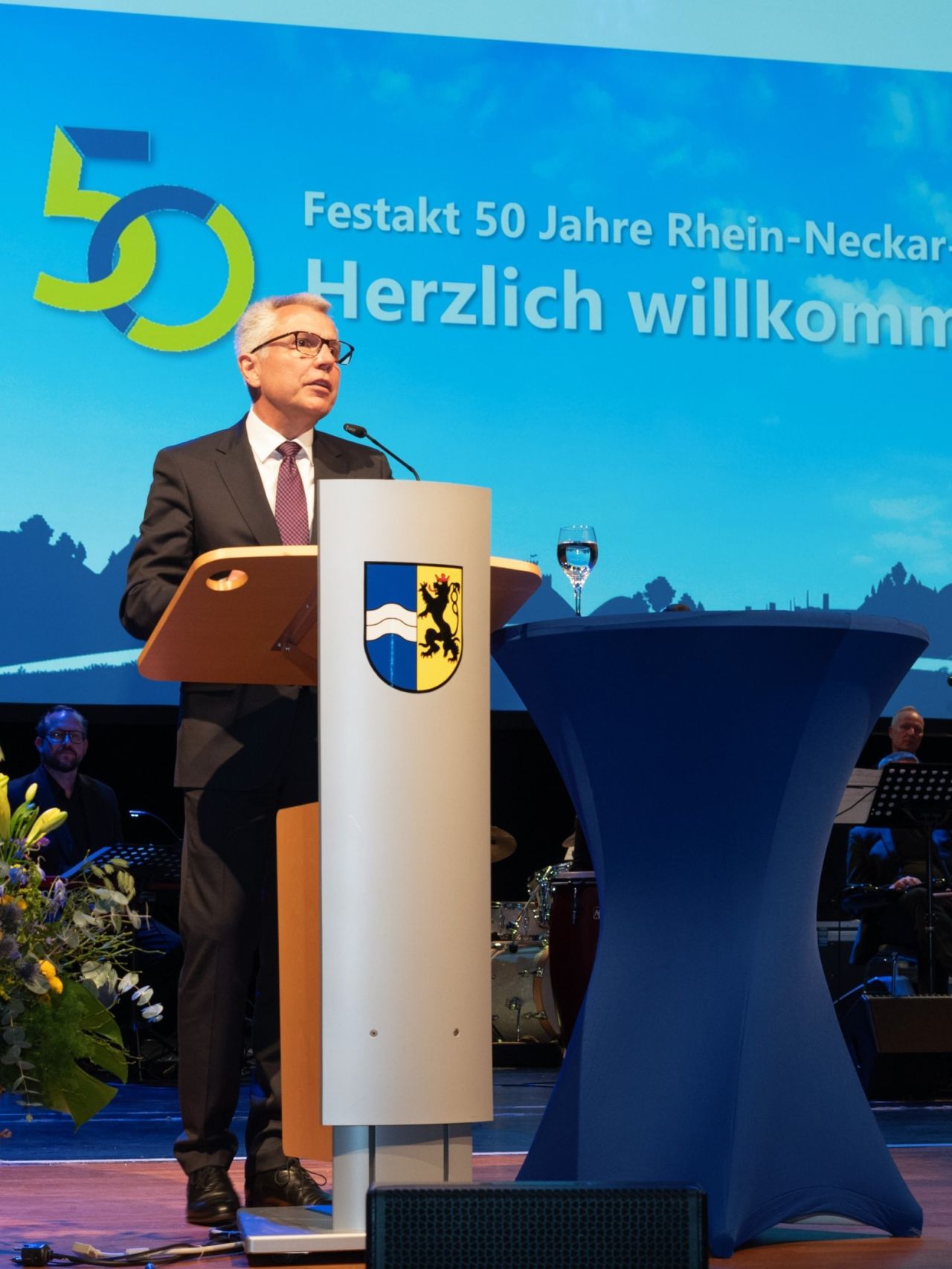 Dieser Alternativtext wurde KI-generiert: Eine Person steht an einem Pult und spricht in ein Mikrofon. Im Hintergrund ist ein Banner mit einer Zahl und Text zu sehen.